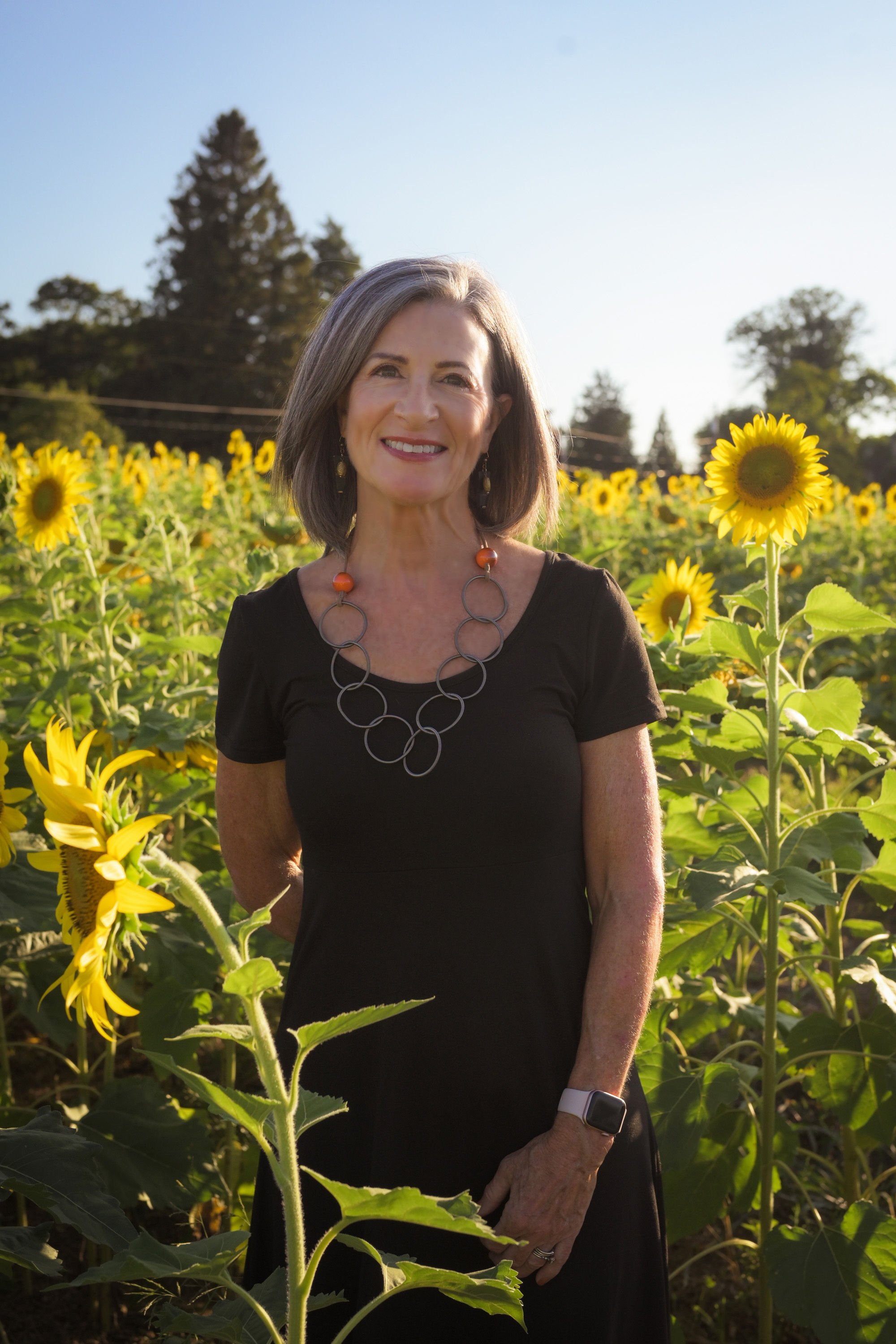 A smiling woman with gray hair and a black dress standing among sunflowers in a field, wearing a circular necklace and a smartwatch.