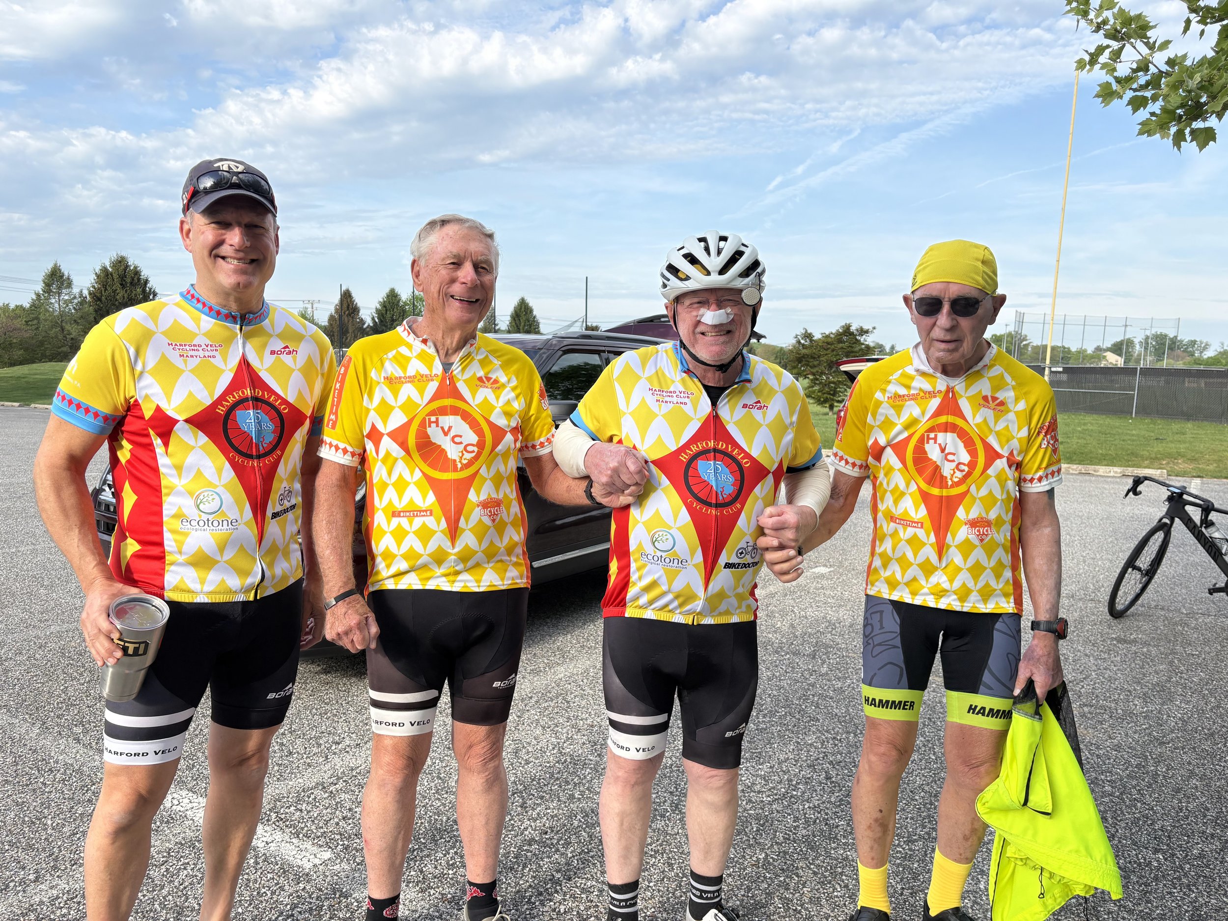 Four older men in cycling gear stand together outdoors, smiling, with a car and a bicycle in the background, under a partly cloudy sky.
