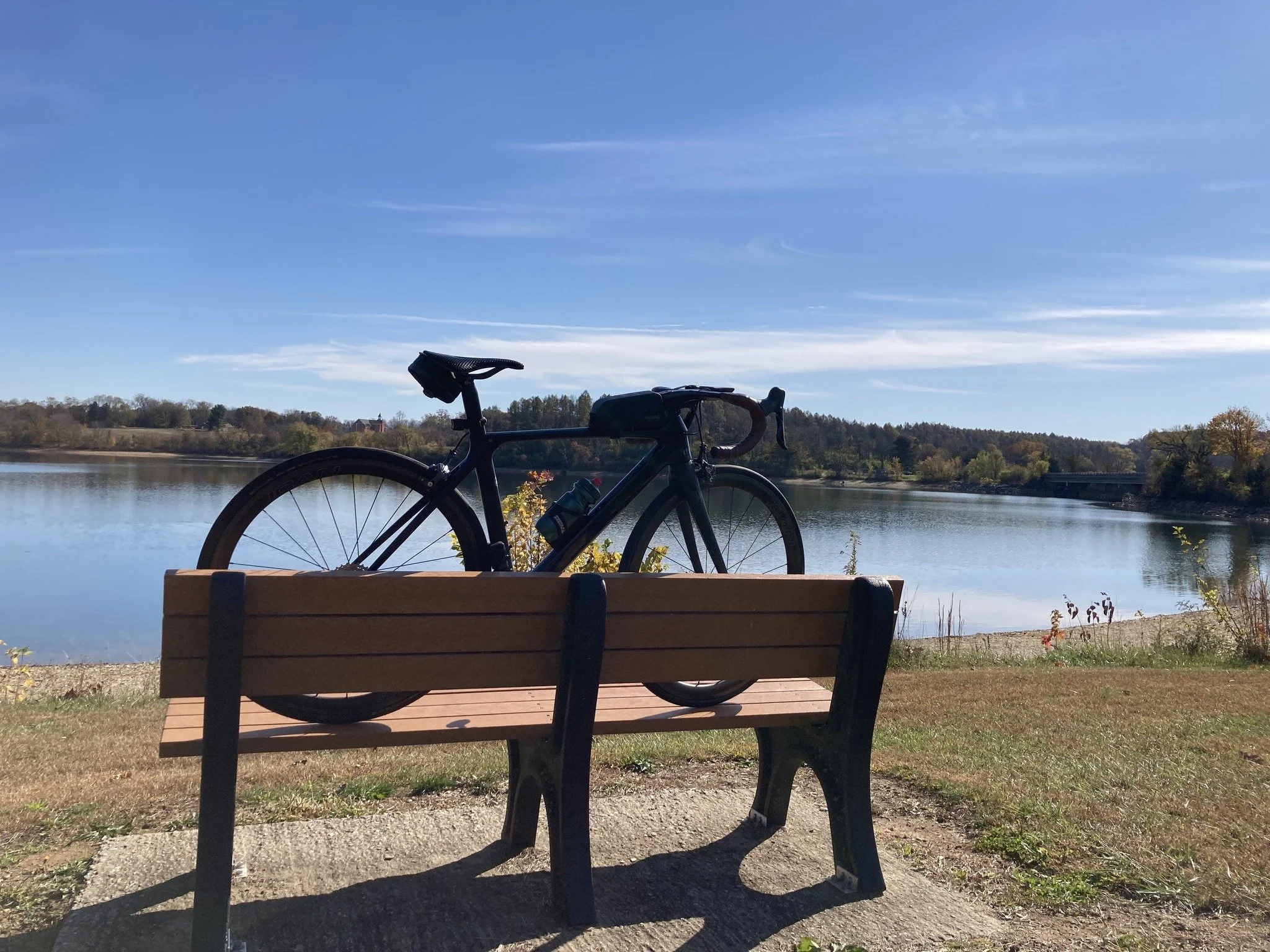 A black bicycle resting against a wooden park bench by a lake on a clear, sunny day with blue skies and distant trees along the shoreline.