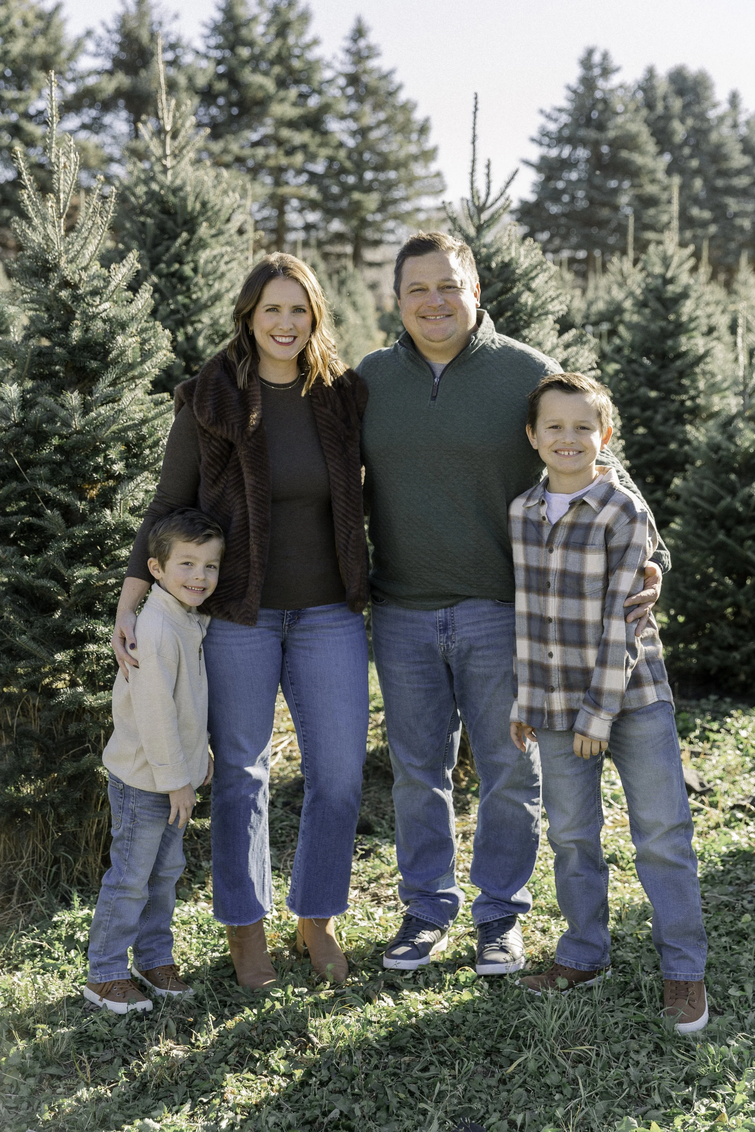 A family of four standing together outdoors among Christmas trees, smiling at the camera.