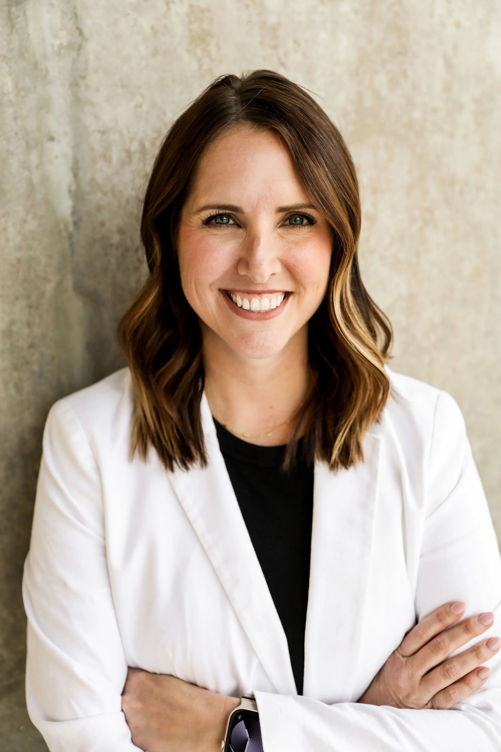 A woman with shoulder-length brown hair and green eyes, smiling, wearing a white blazer over a black top, standing against a textured beige wall.