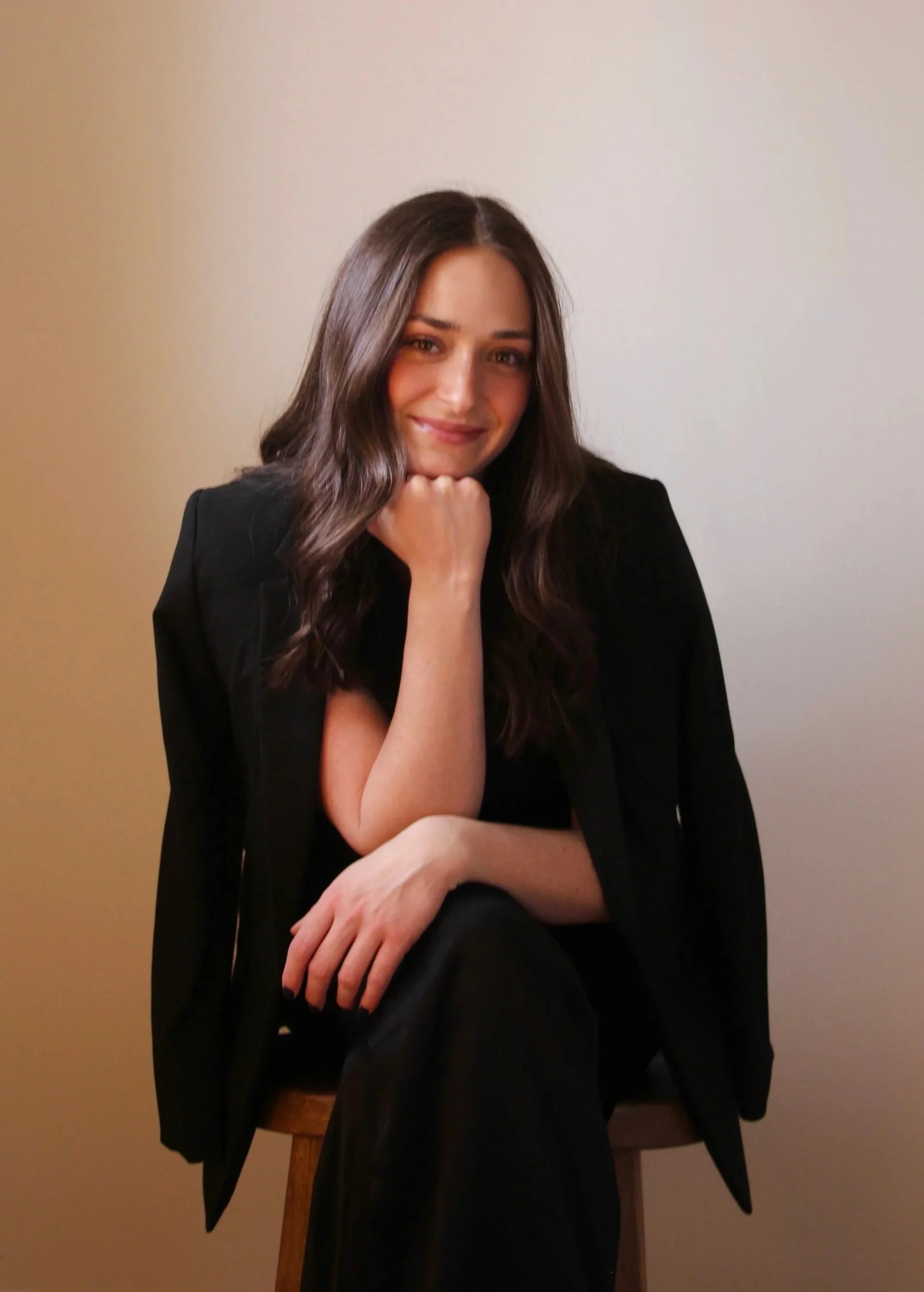 A young woman with long brown hair sitting on a wooden chair against a plain light-colored wall. She is wearing a black blazer and smiling softly, resting her chin on her left hand with her right arm crossed over her lap.
