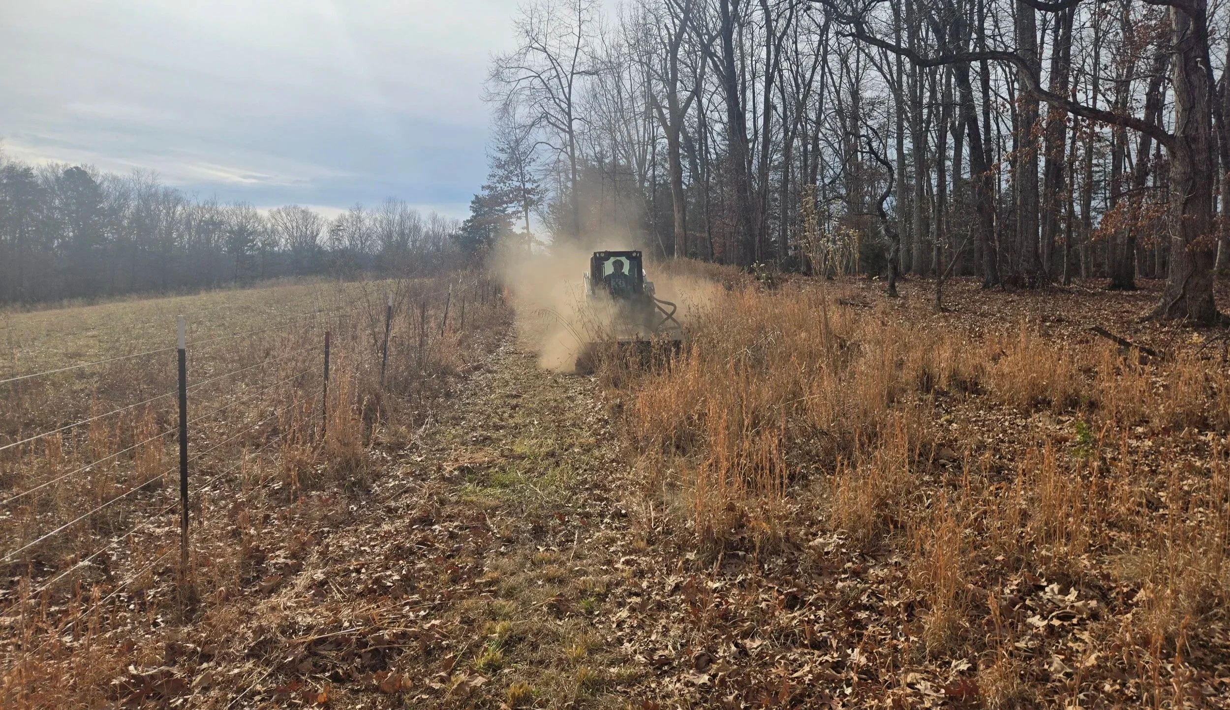 A tractor working on a hillside with dry grass and leafless trees in the background, near a wire fence on the left side of the image.