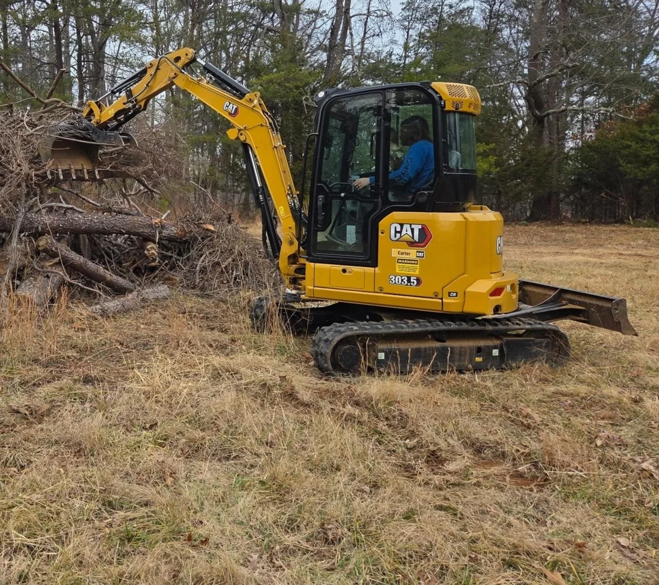 A yellow compact excavator is digging into a pile of fallen tree branches and logs in a grassy field with trees in the background.