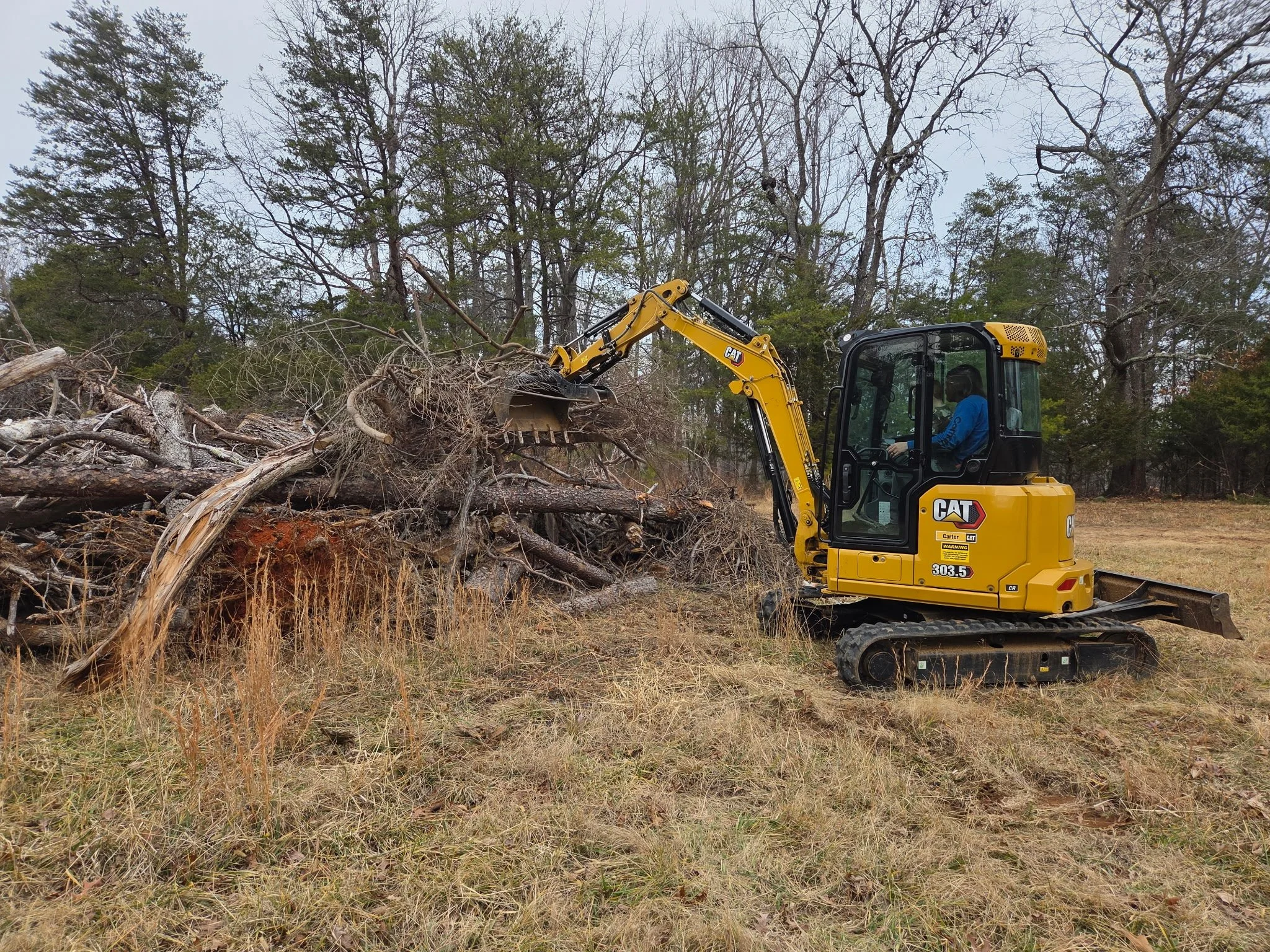 A mini excavator is clearing a pile of fallen tree branches and logs on a grassy field