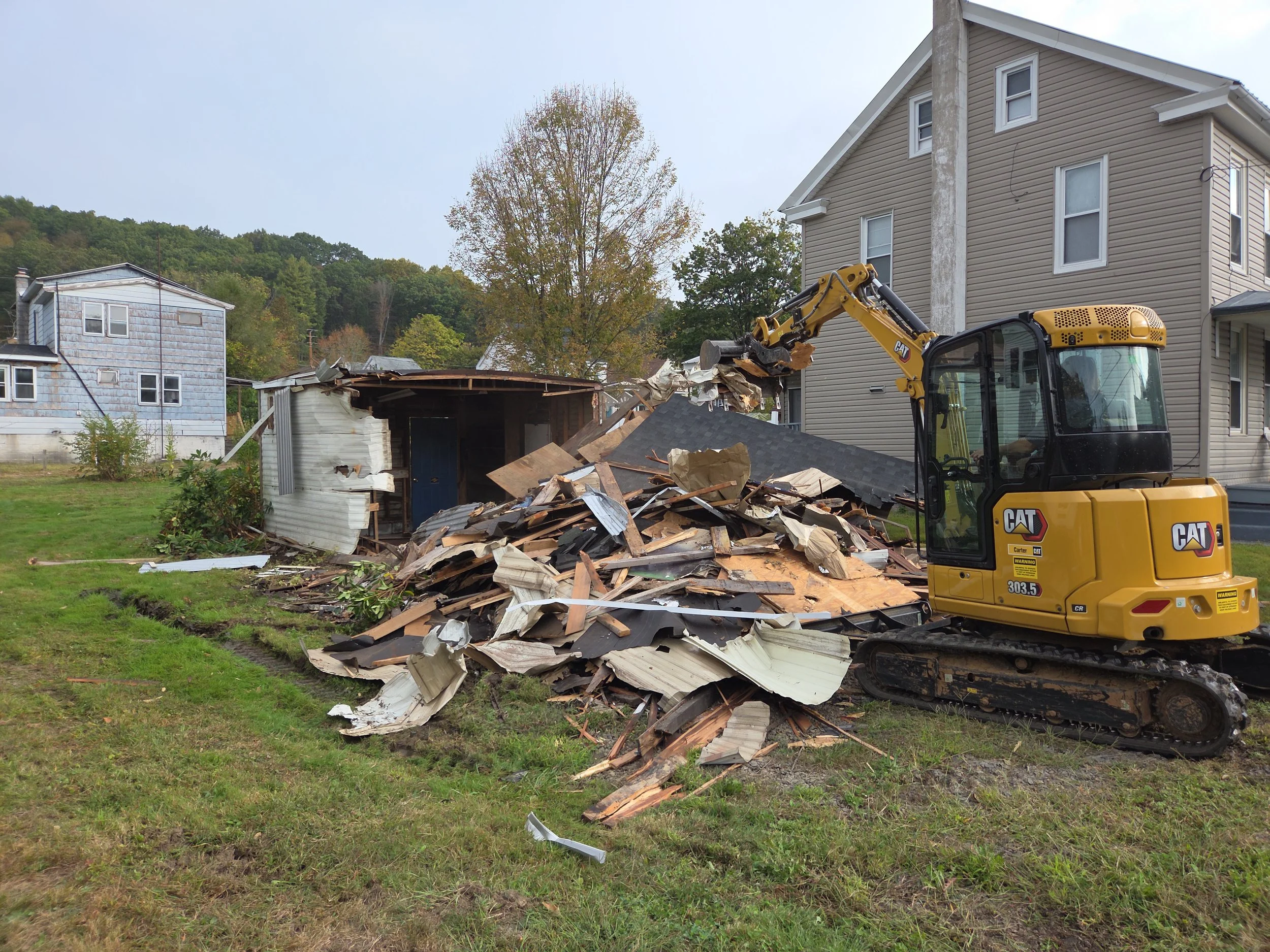 A yellow CAT excavator demolishing a small wooden building, with debris and broken wooden panels scattered on the ground, near residential houses and trees.