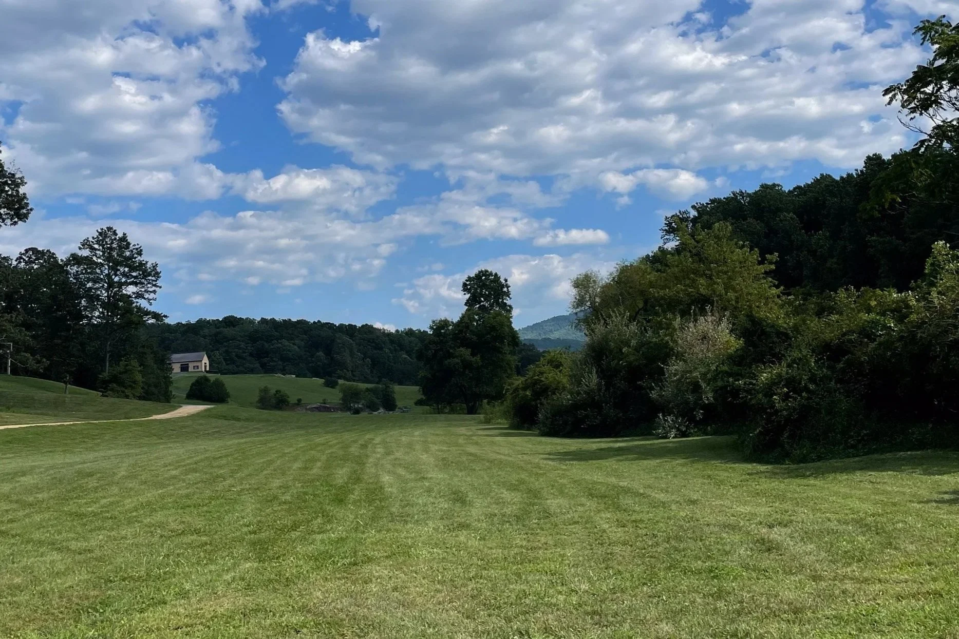 A scenic landscape with a well-maintained grassy field, trees on either side, a small building in the distance, and a partly cloudy sky overhead.