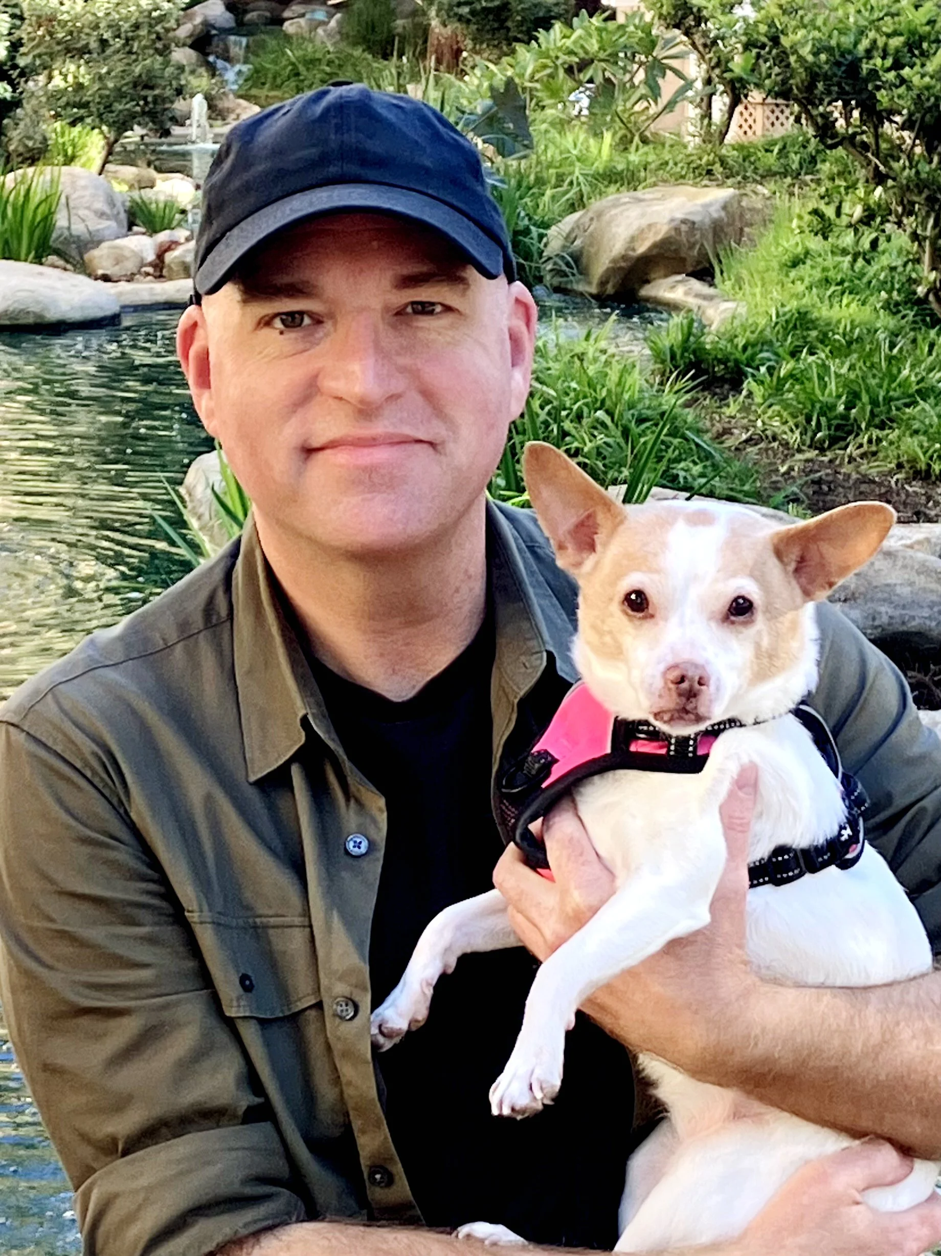 A man in a black cap and khaki shirt holding a small light-colored dog with pointy ears, near a pond with rocks and green plants in the background.