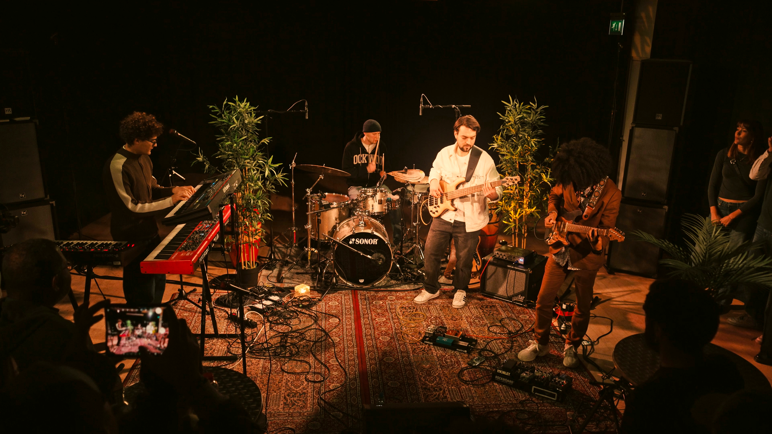 A band performing live on stage with four members playing keyboards, drums, bass, and guitar, surrounded by large potted plants, with audience members watching and one person recording with a smartphone.