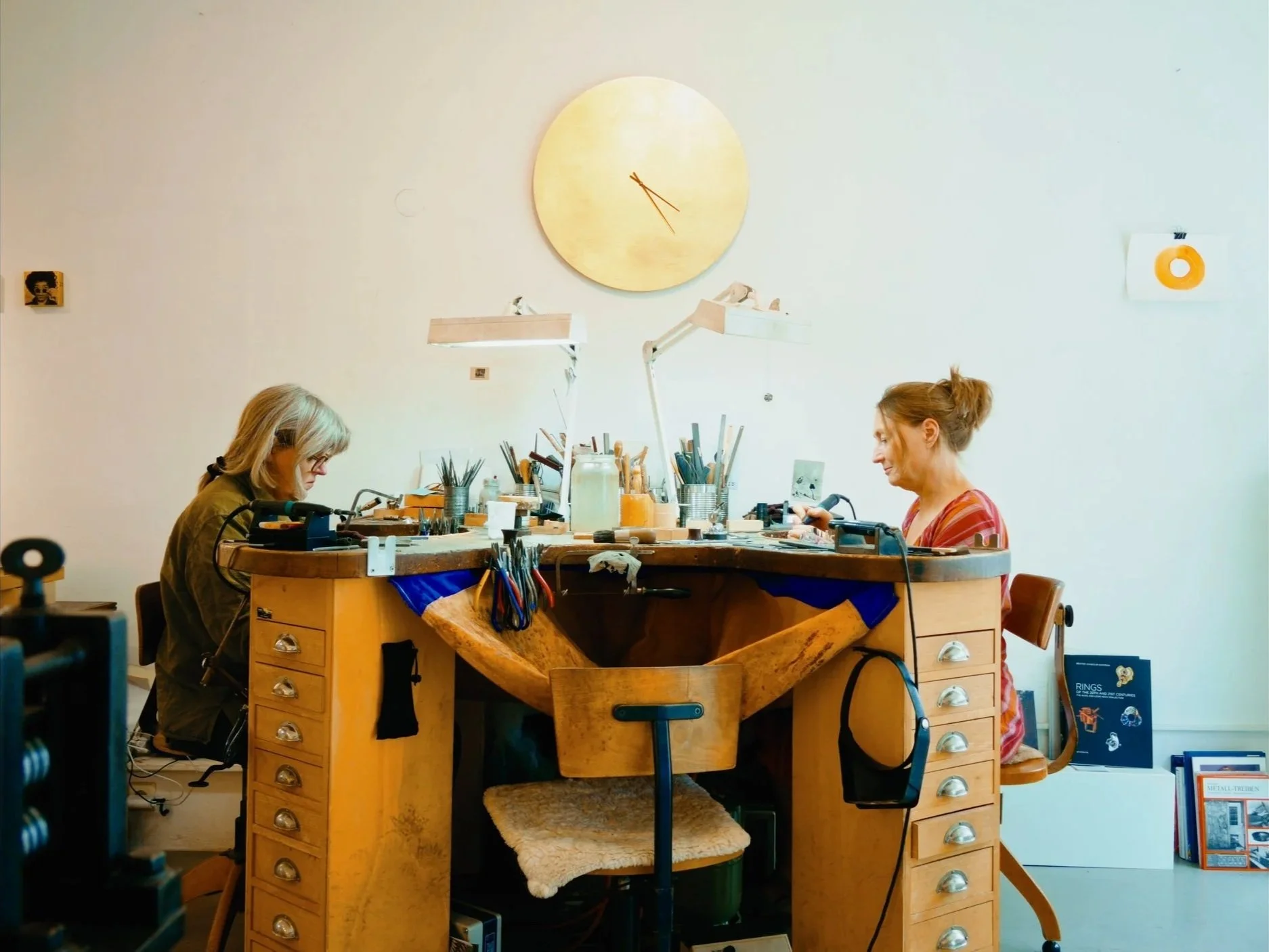 Two women working at a large wooden jewelry polishing and repair station in a well-lit studio surrounded by tools, with a large clock on the wall behind them.