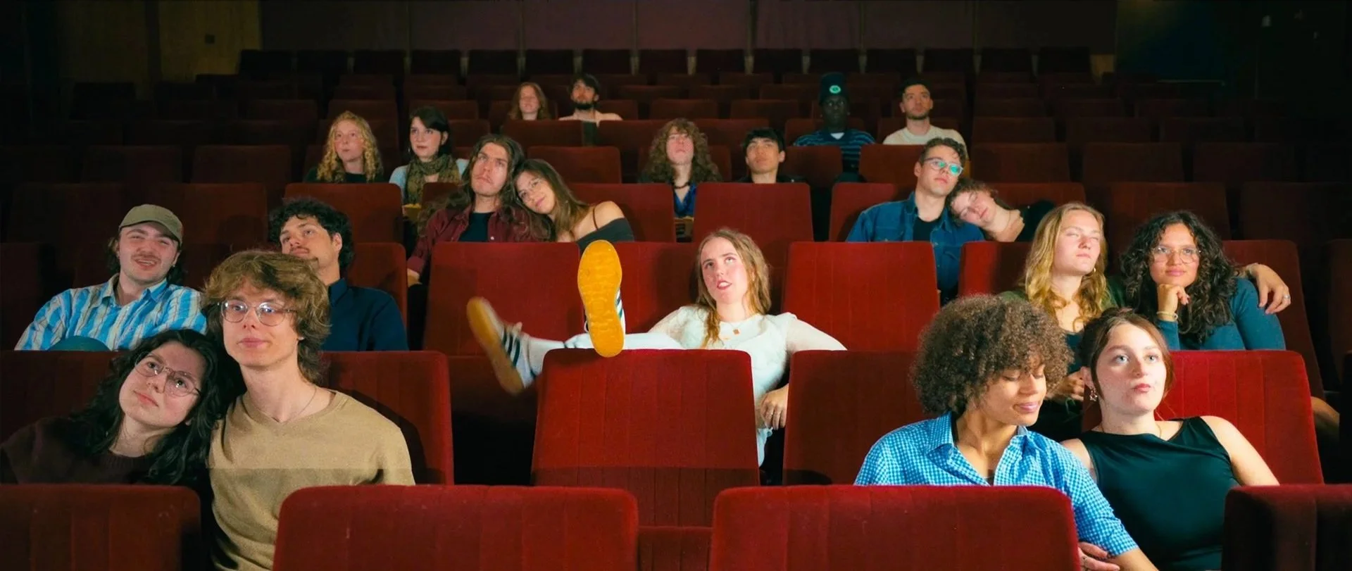 A group of people sitting in a darkened theater or cinema, with some appearing sleepy or bored, and one person in the center wearing pajamas with their legs up on the seat.