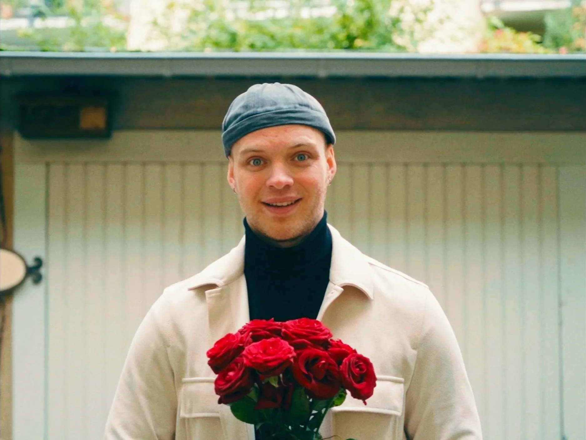 A smiling young man wearing a gray beanie and black turtleneck, holding a bouquet of red roses, standing outdoors near a wooden structure.