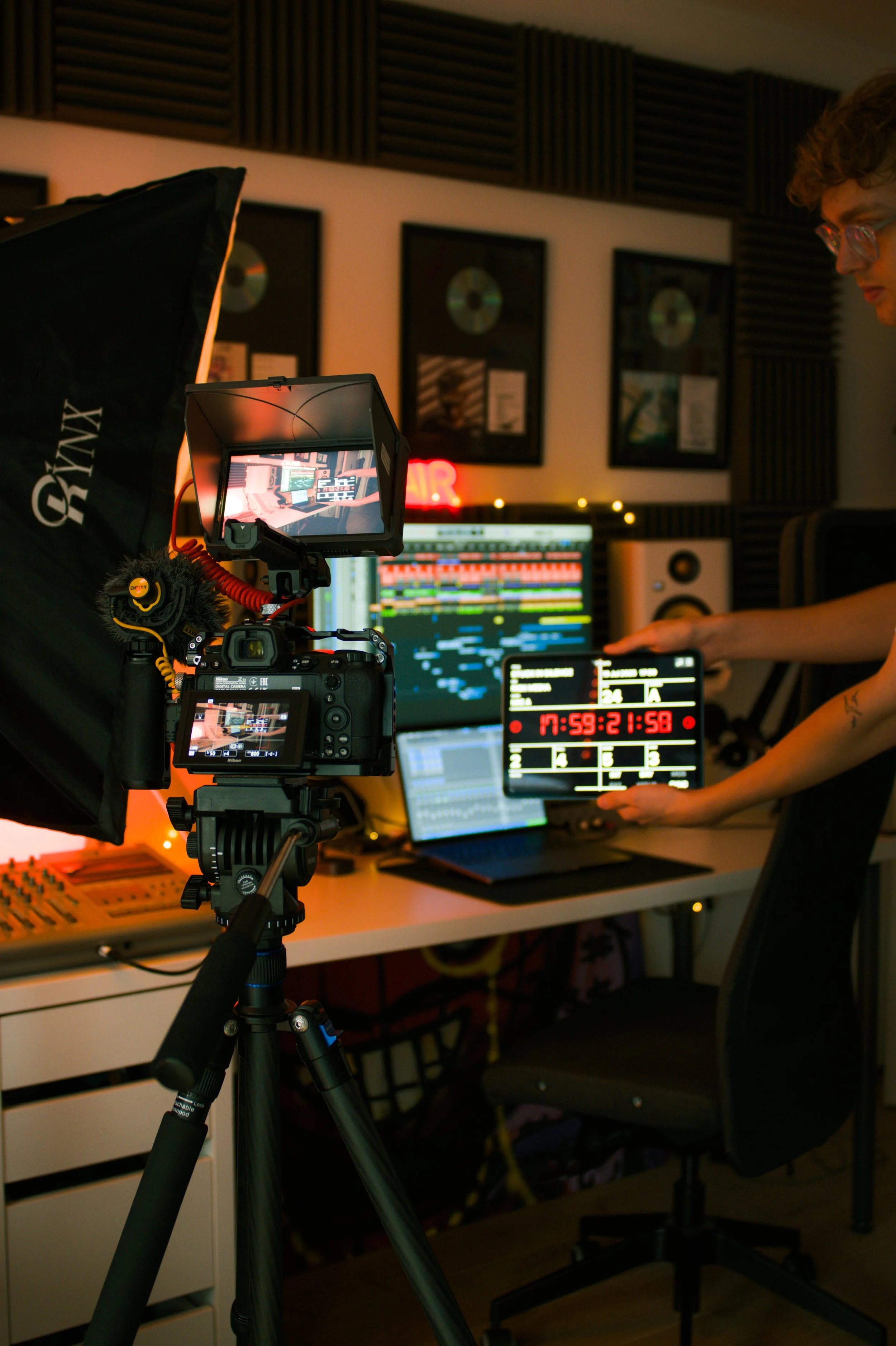 A person is working on music production with a camera recording the process in a home studio. The setup includes monitors, computers, and soundproofing on the walls.