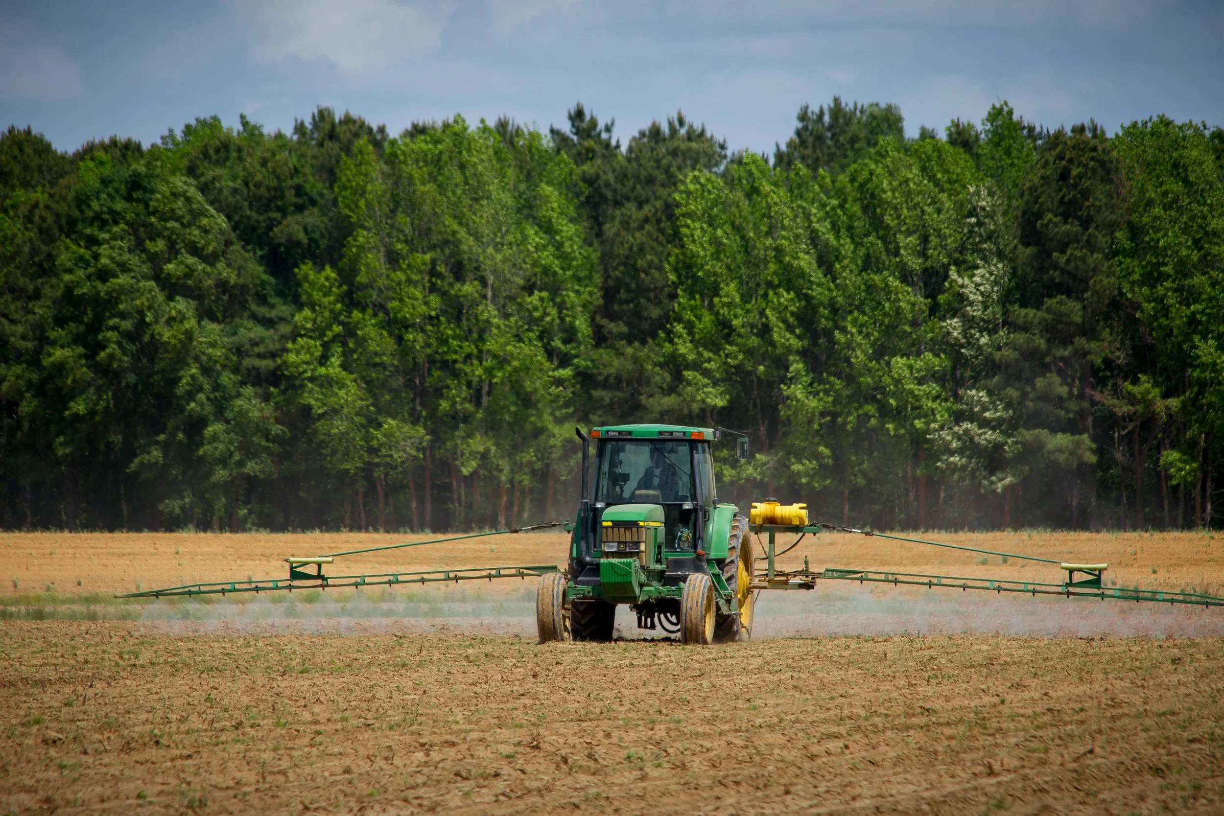 A green tractor with a yellow tank is spraying a field with a liquid, with a backdrop of green trees and a partly cloudy sky.