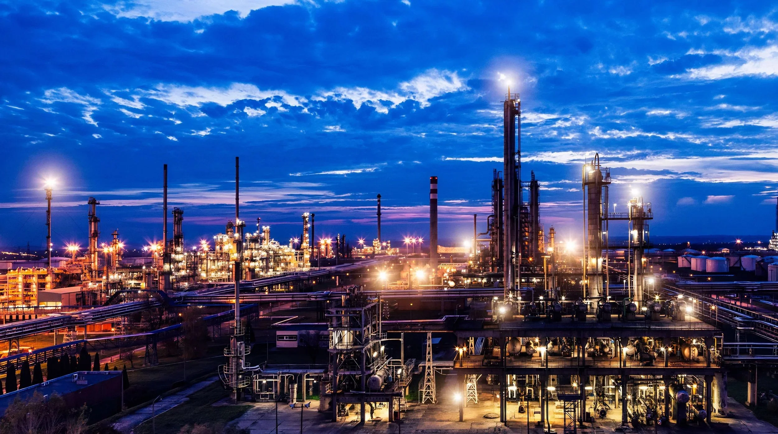 A large industrial oil refinery at dusk with illuminated pipes, tanks, and tall processing towers against a partly cloudy sky.