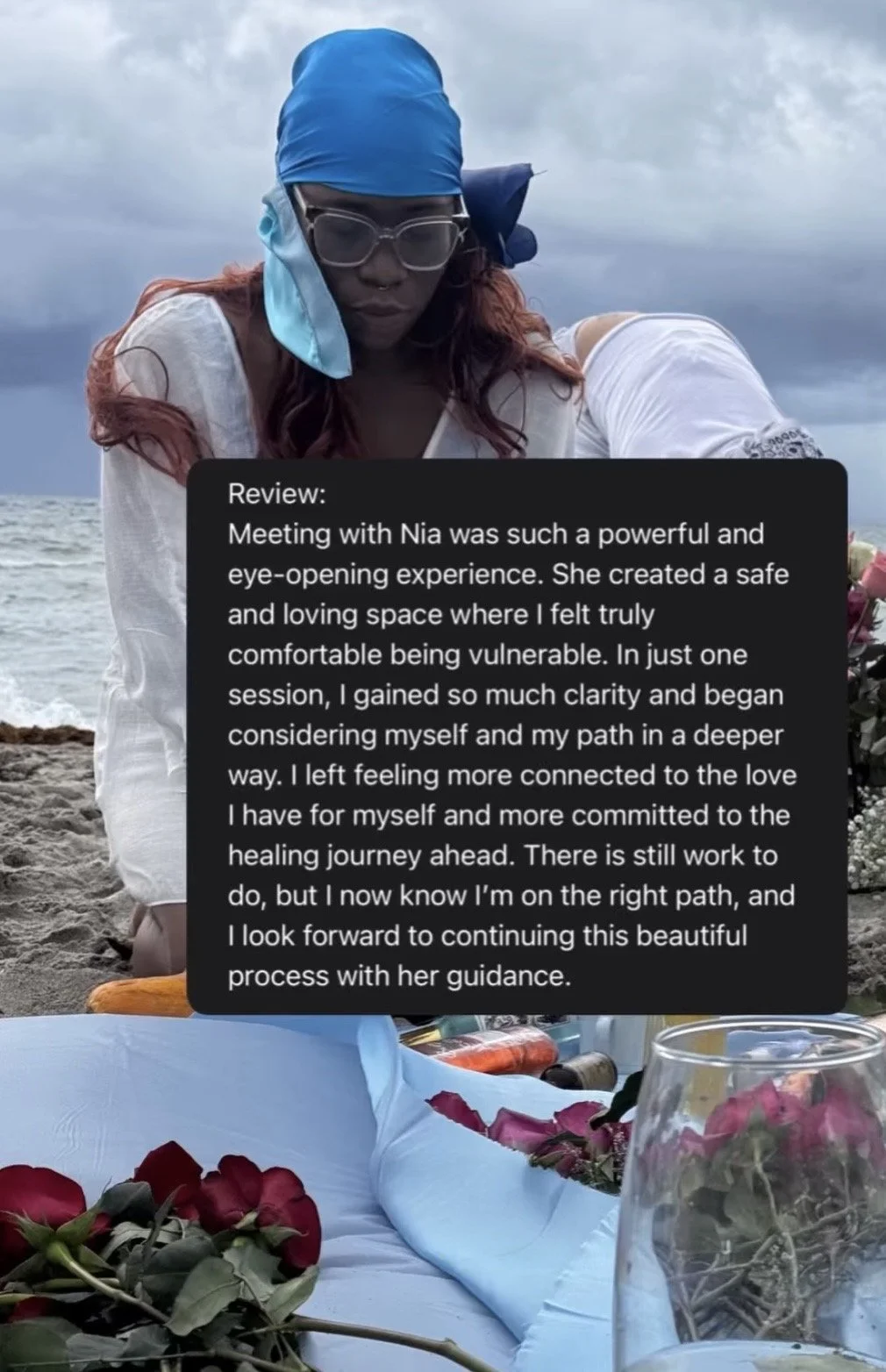 Woman with red hair, wearing glasses, a blue headscarf, and white clothing, sitting on a sandy beach with an ocean and cloudy sky in the background. In front of her, there is a table with flowers, a wine glass, and bottles.