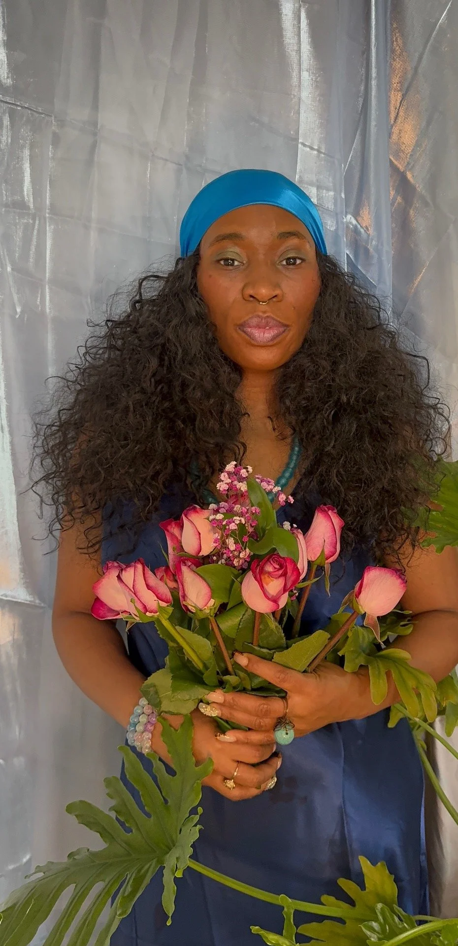 A woman with long curly hair wearing a blue headscarf and jewelry, holding a bouquet of pink roses and greenery, standing in front of a silver background.