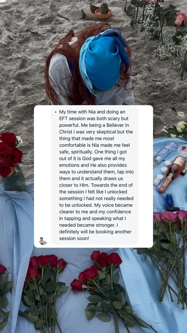 A person with long red hair and a blue headscarf kneeling on sand next to a memorial site with flowers and candles.