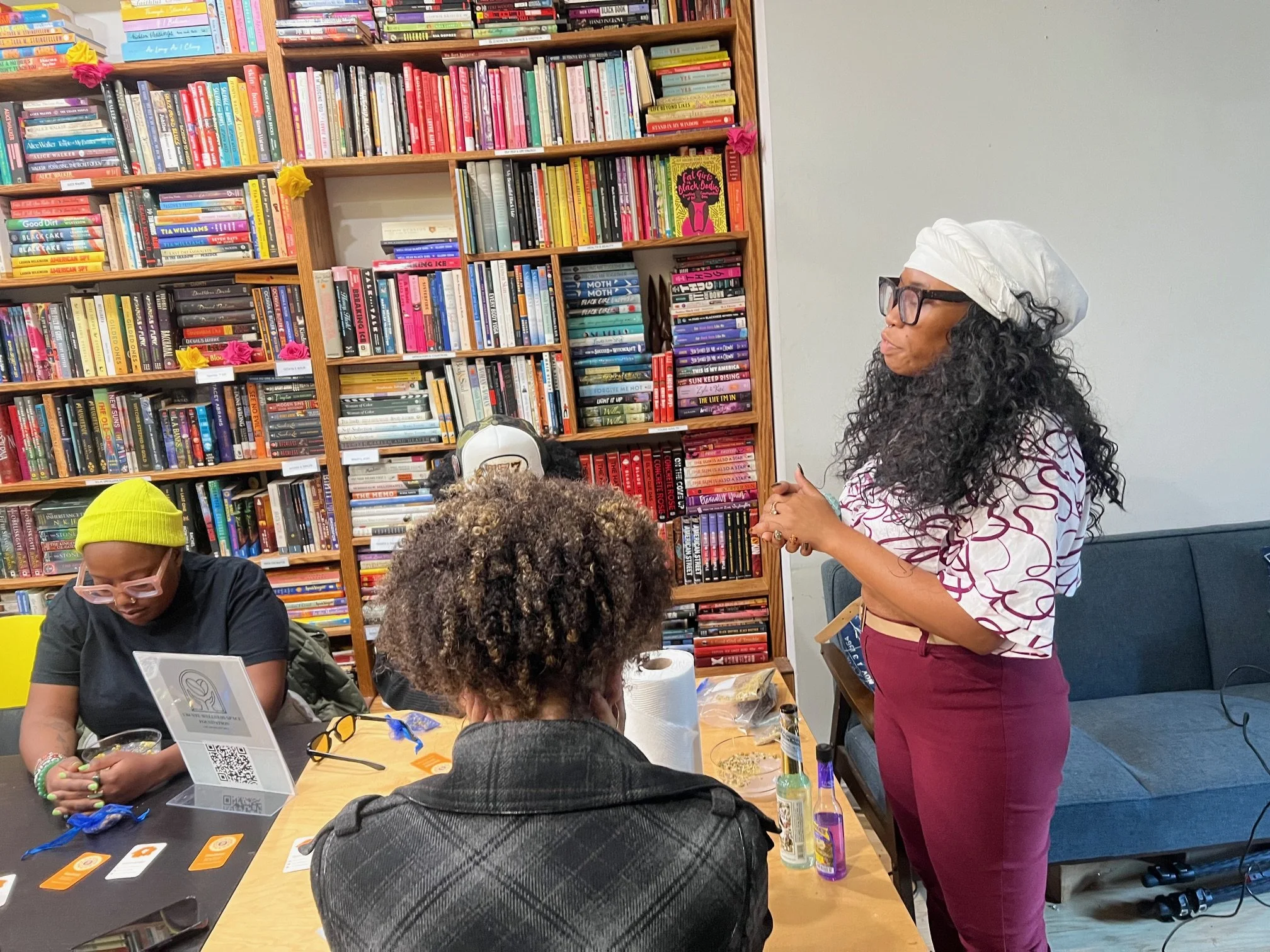 A woman wearing glasses, a white headscarf, and patterned shirt stands in front of a bookshelf, speaking to a group in a room with other women sitting at a table surrounded by various items, including bottles, cards, and a QR code.