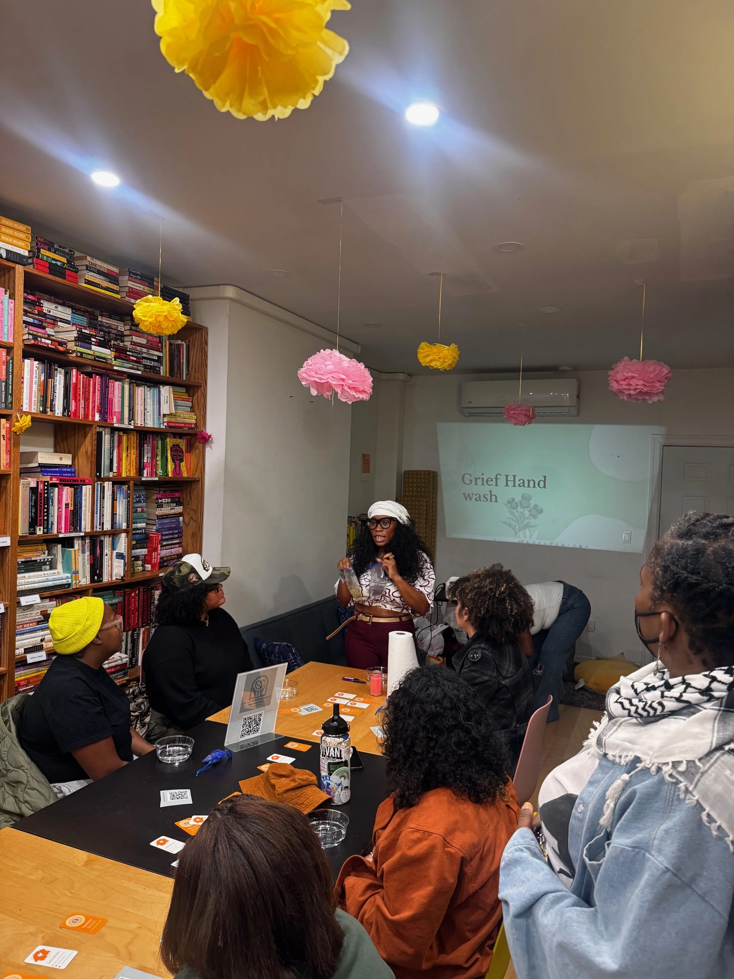 A woman stands in front of a room of seated people giving a presentation about grief hand wash. The room has bookshelves, pink and yellow paper decorations hanging from the ceiling, and a projector screen displaying the presentation.