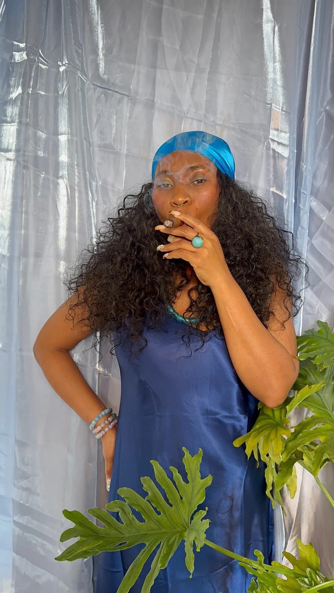 A woman with curly hair wearing a blue headscarf and dress, holding a cigar, standing in front of a shiny silver curtain with green plants in the foreground.