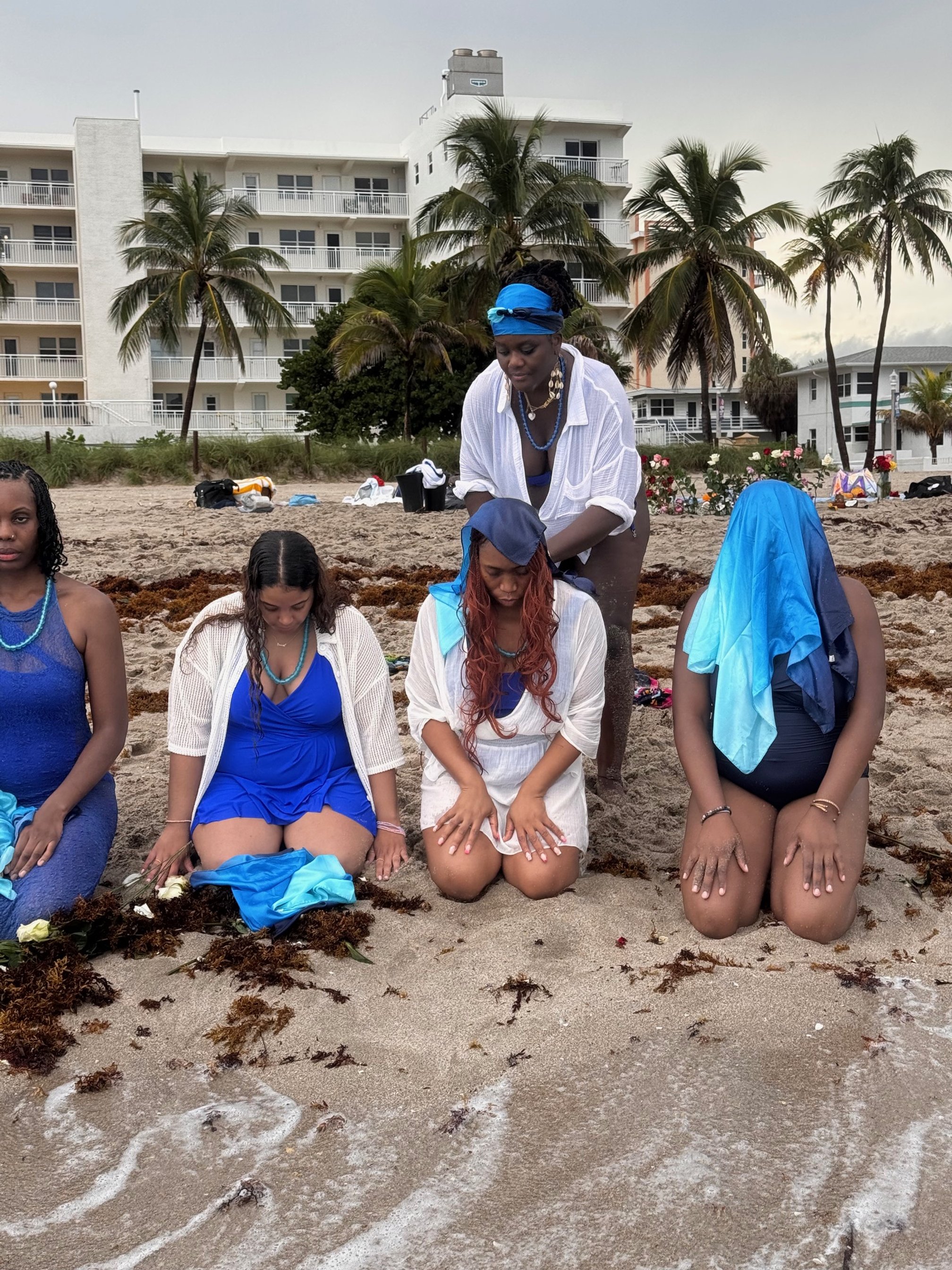 Four women kneeling on the sand at the beach, with a woman standing behind them, participating in a ritual or ceremony involving tying cloth and flowers to the sand, with palm trees and buildings in the background.