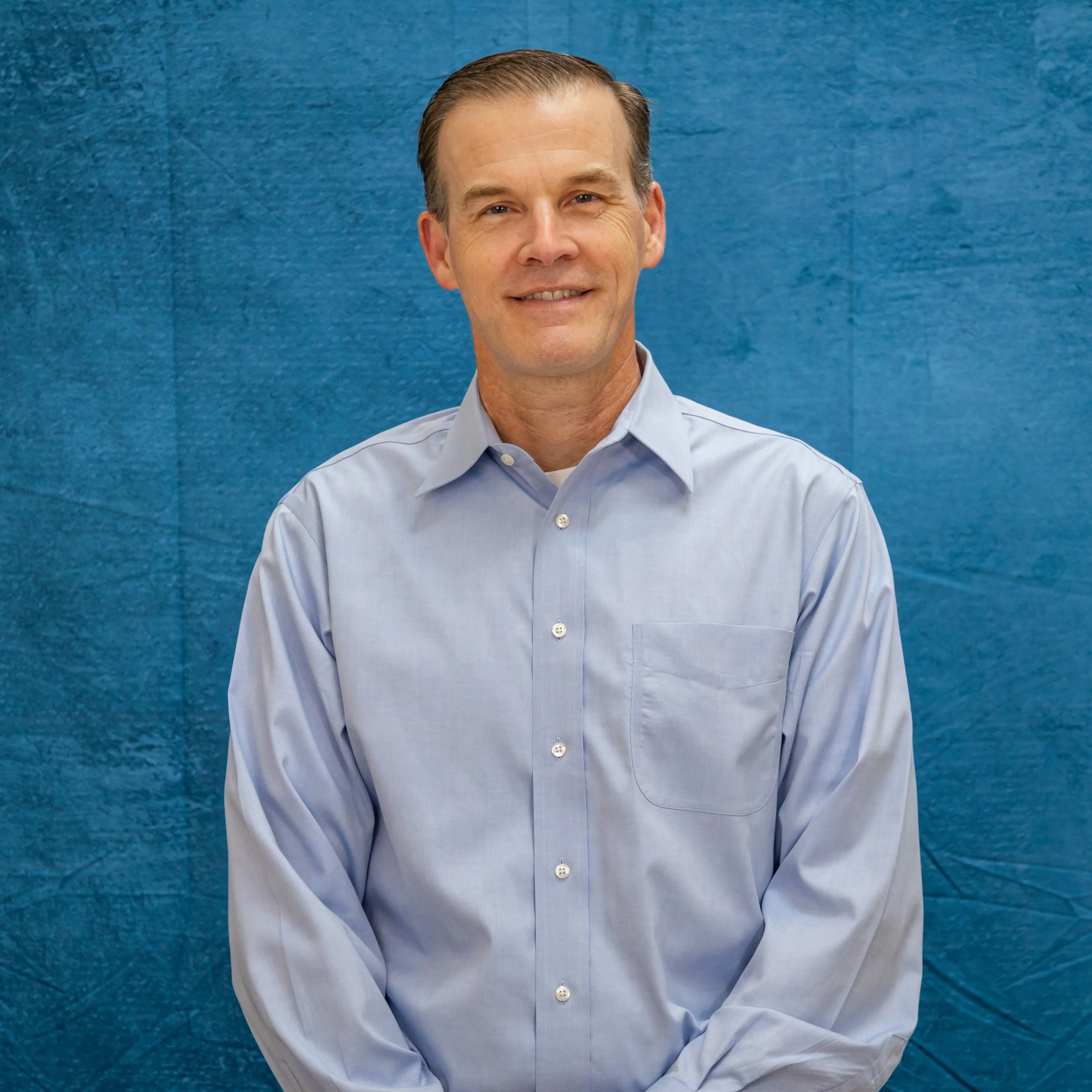 A middle-aged man with neatly combed brown hair, wearing a light blue dress shirt, smiling against a blue textured wall background.