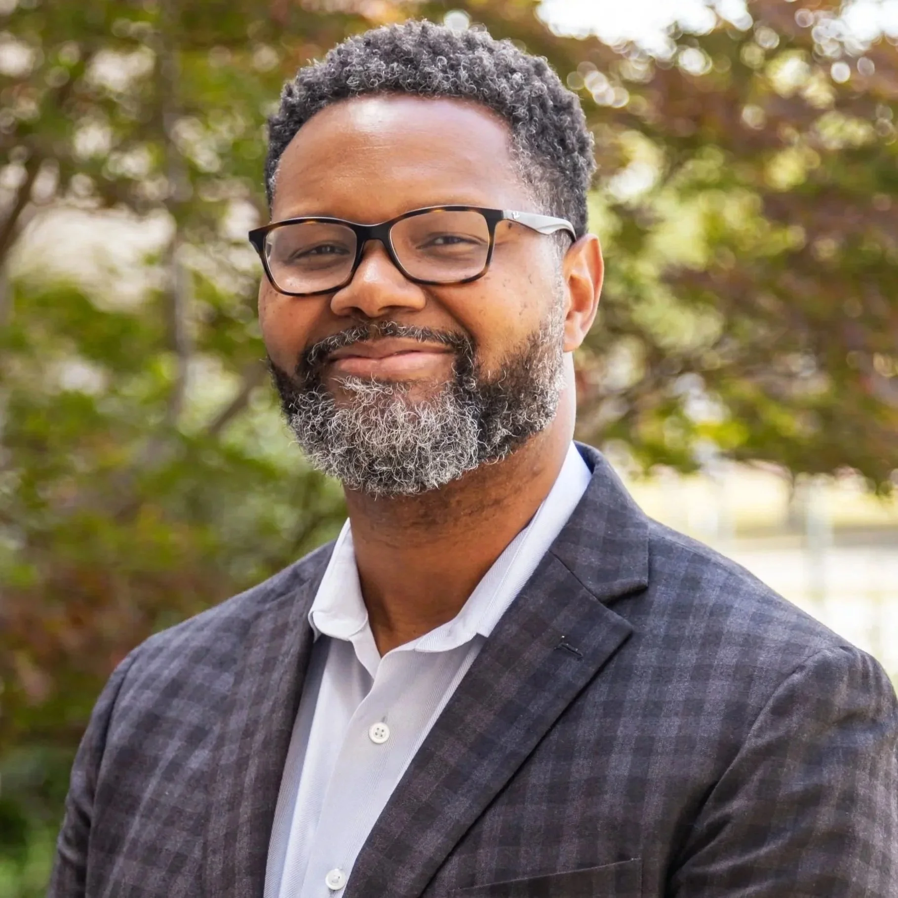 Close-up of a smiling man wearing glasses and a gray plaid suit, standing outdoors with blurred autumn-colored trees in the background.