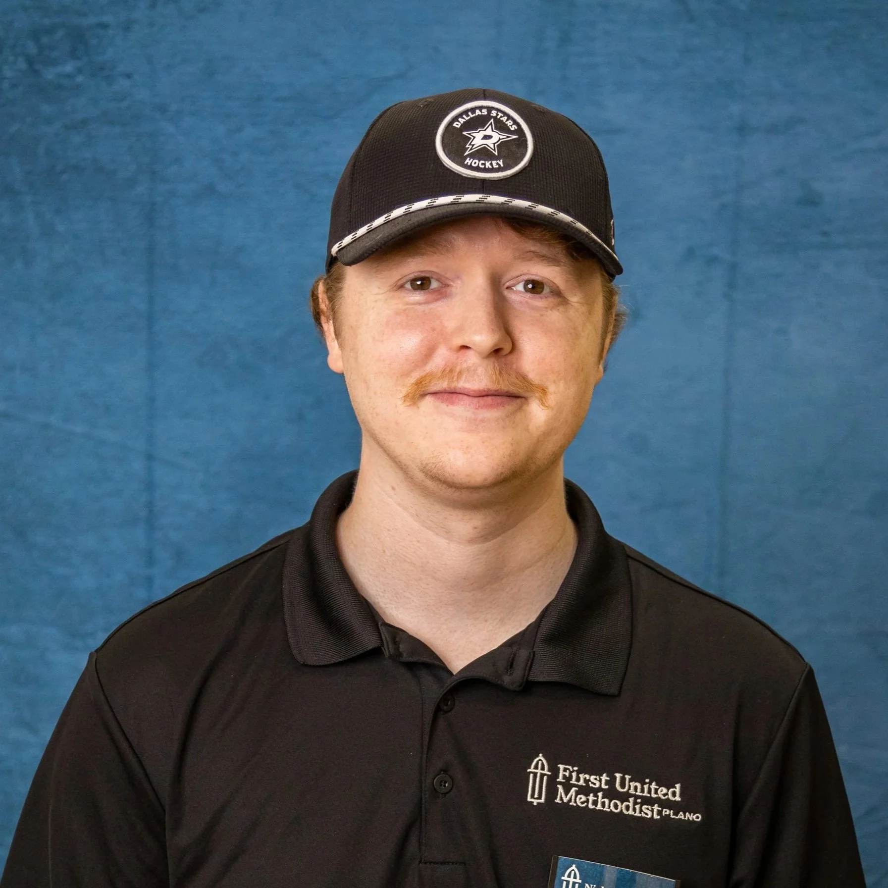 A man wearing a black polo shirt with a logo that reads 'First United Methodist Plano' and a black baseball cap with a Dallas Stars hockey team logo against a blue background.