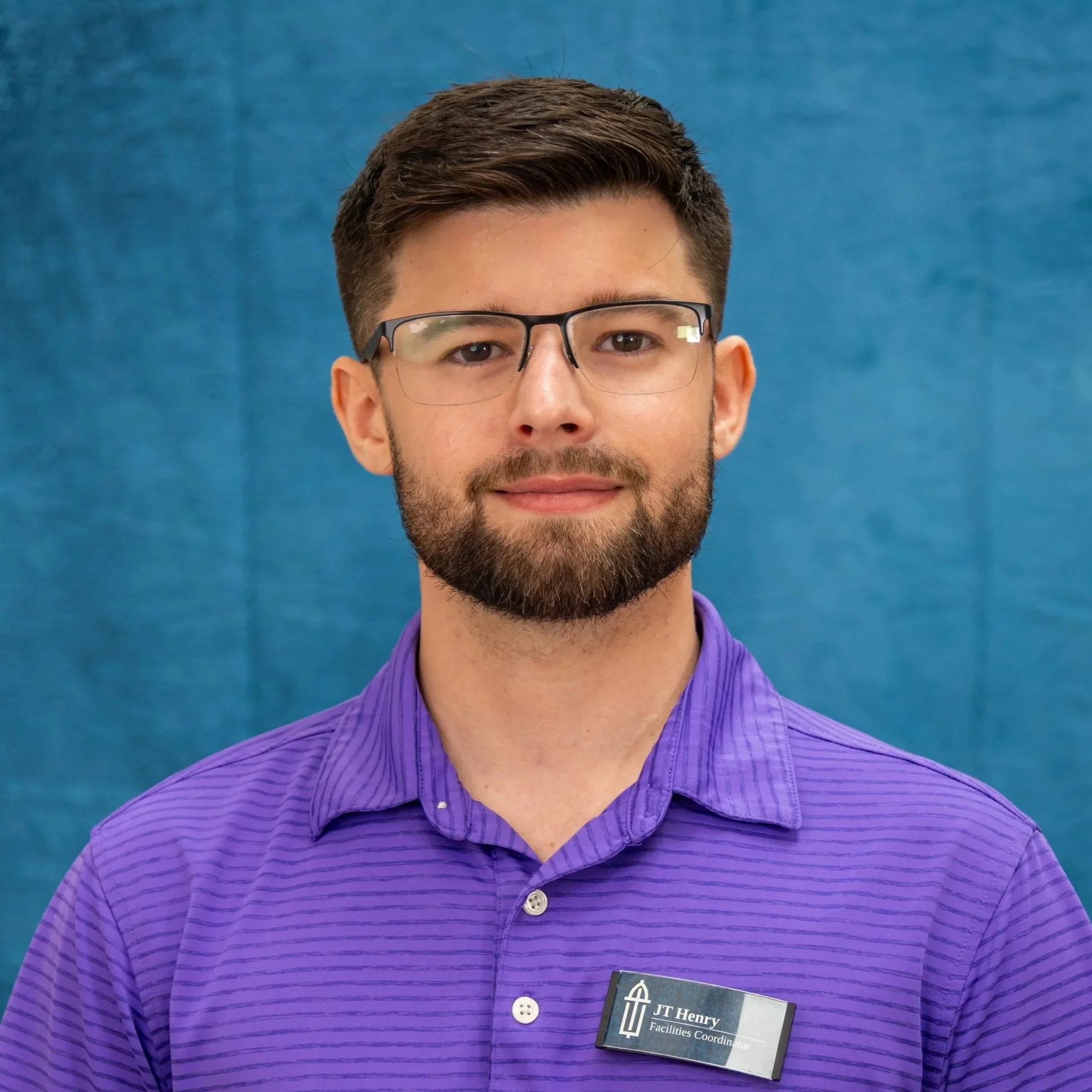 A man with brown hair, glasses, and a beard wearing a purple striped shirt and a name tag that reads "JT Henry, Facilities Coordinator," stands against a blue background.