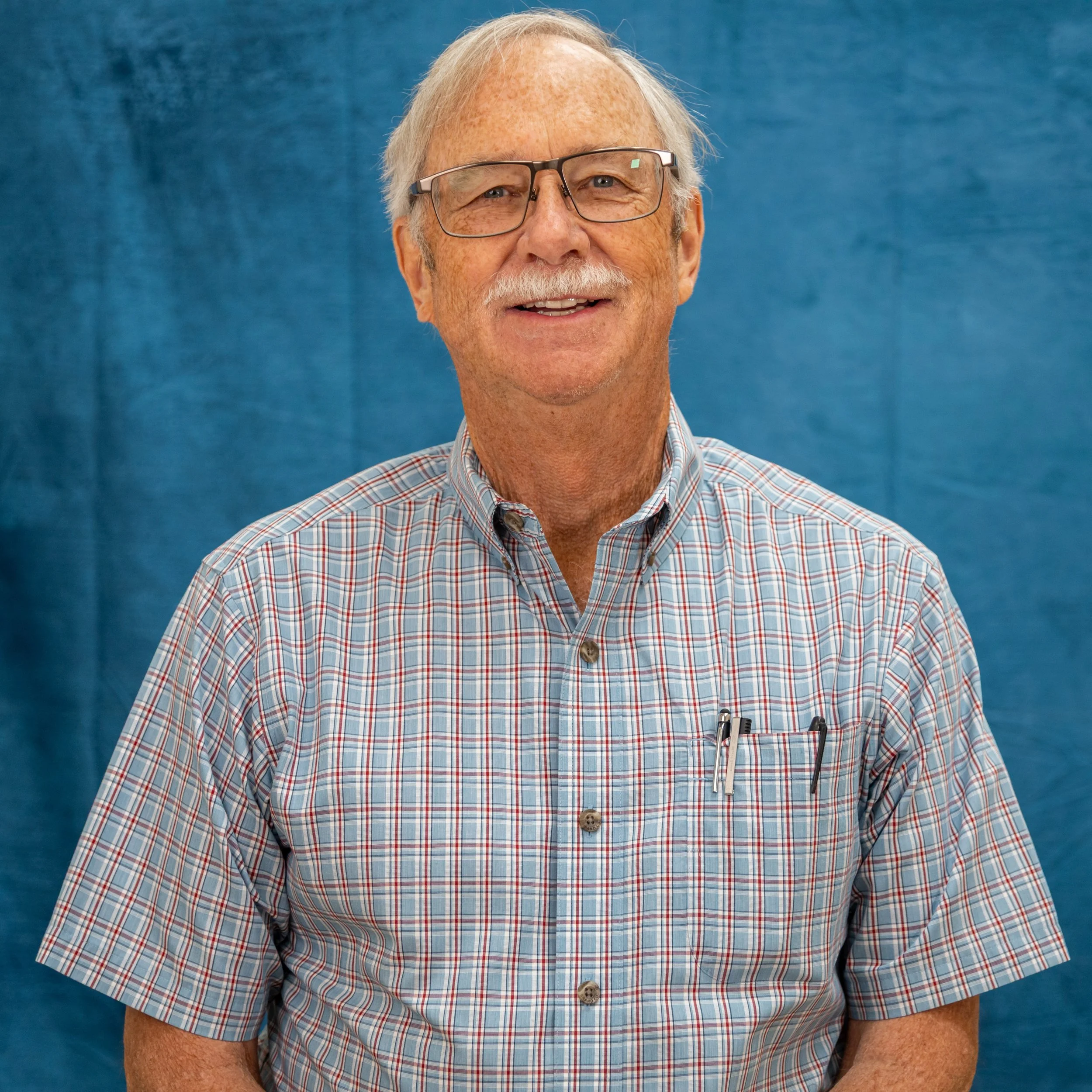 A smiling elderly man with white hair and glasses, wearing a checkered shirt, standing in front of a blue background.