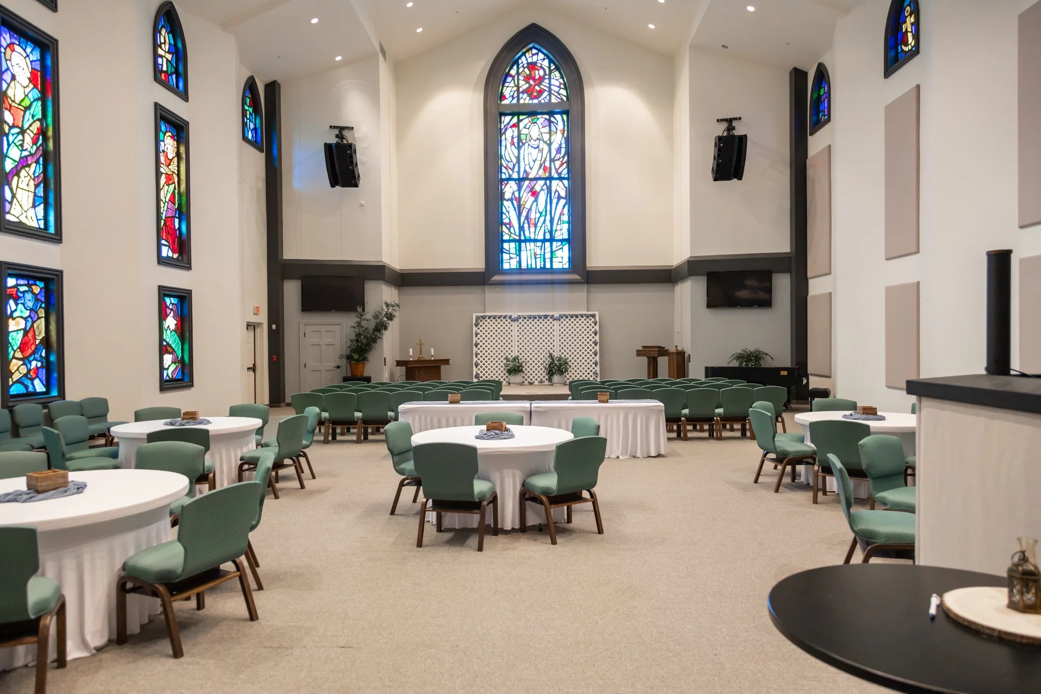 Interior of a church or chapel with green chairs, round tables with white tablecloths, stained glass windows, a small altar with a cross, plants, and two large speakers mounted on the walls.