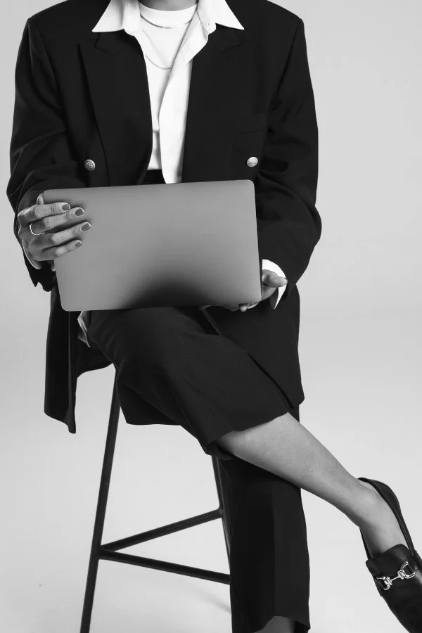A person sitting on a chair, dressed in a black suit with a white shirt, working on a laptop in a studio with a plain background.