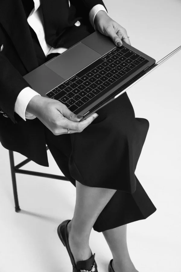 A woman dressed in a black blazer and skirt sitting on a chair, holding a closed laptop on her lap, with her legs crossed.