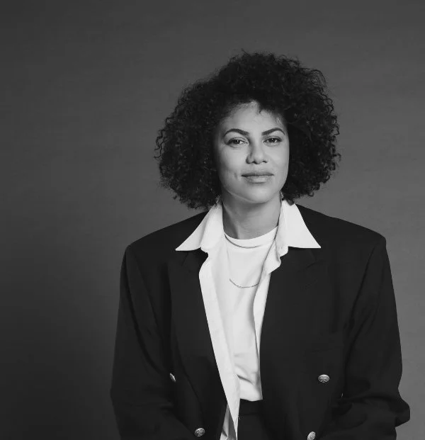 Portrait of a woman with curly hair wearing a blazer over a white top against a plain background.
