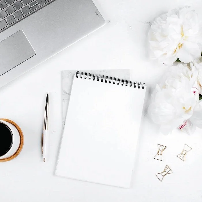 A white desk with a silver laptop in the top left corner, a spiral notebook with blank pages in the center, a black and silver pen to the left of the notebook, a cup of coffee in a wooden holder to the left, a bunch of white flowers on the right, and three gold paper clips near the flowers.