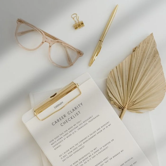 Flat lay of pink eyeglasses, a gold pen, a gold binder clip, a white clipboard with a career clarity checklist, and a dried palm leaf on a white surface.
