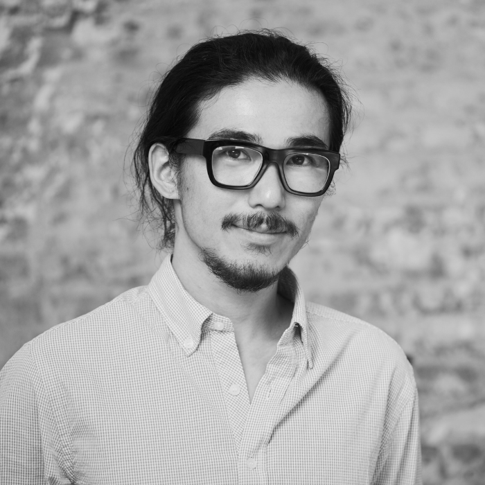 Black and white portrait of a man with glasses and a beard, wearing a collared shirt, in front of a brick wall.