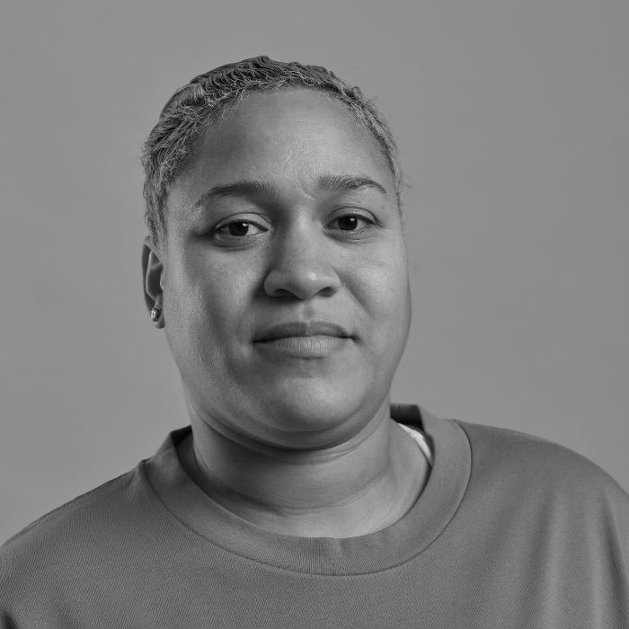 Black and white portrait of a woman with short curly hair, wearing a crewneck shirt and small earrings, looking directly at the camera with a neutral expression.