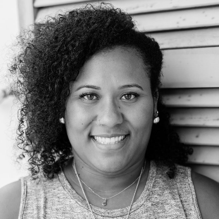A smiling woman with curly hair and a nose ring, wearing a sleeveless shirt and necklaces, standing next to a wooden wall.