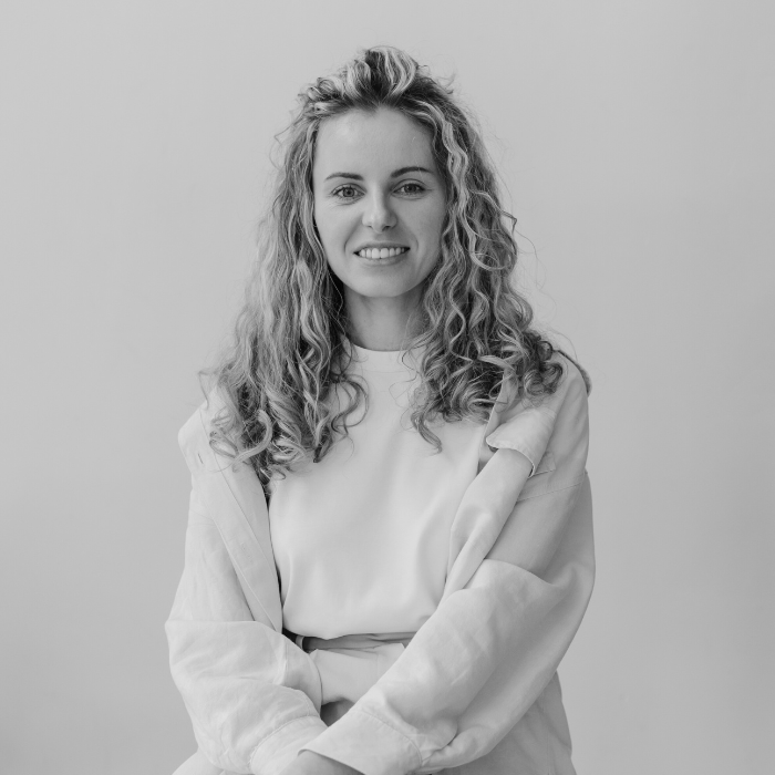 Black and white photo of a young woman with curly hair, smiling, wearing a white t-shirt and a light-colored jacket, standing against a plain background.