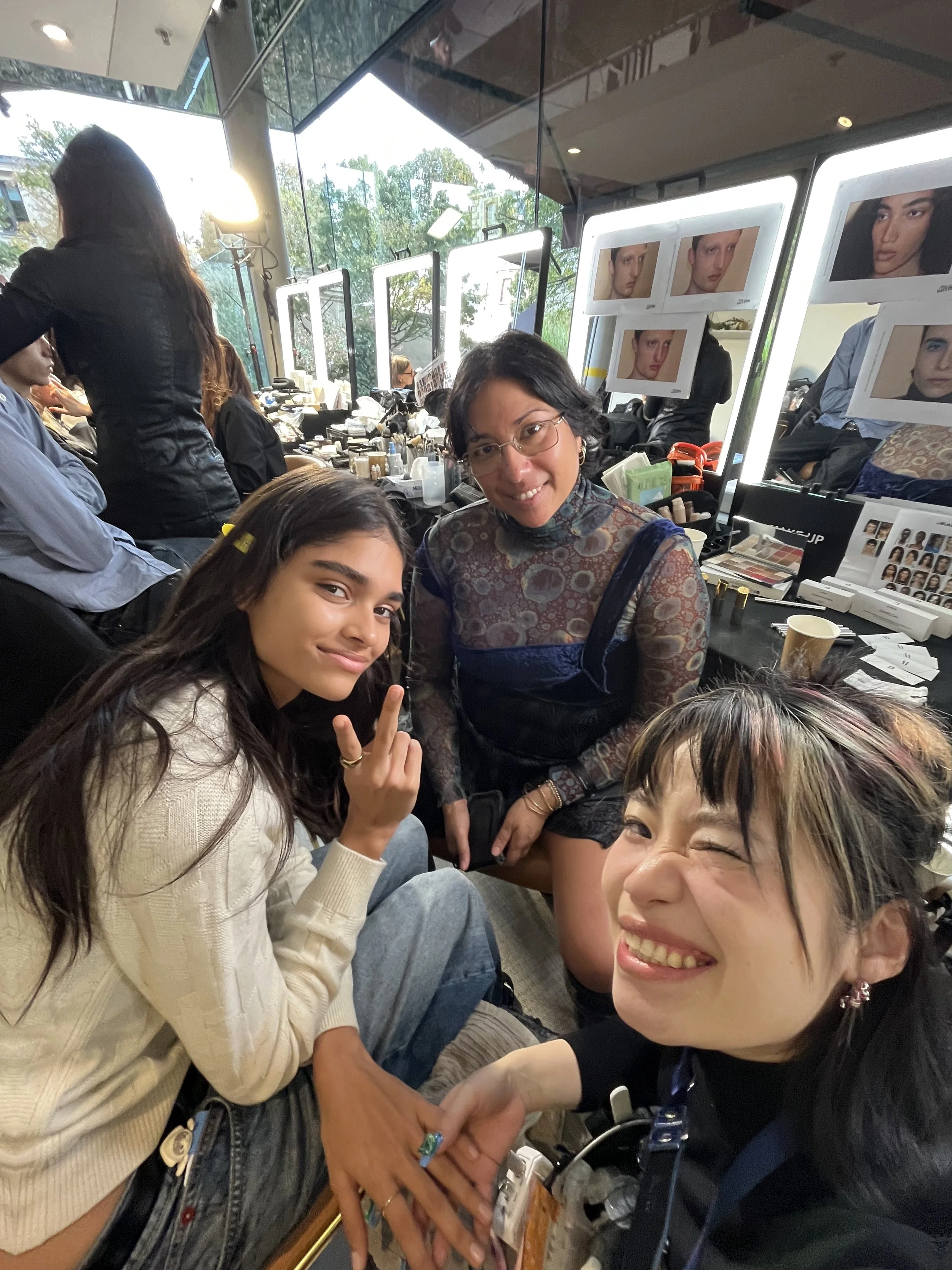 Three women in a makeup room, with photography and makeup tools around, smiling and posing for a selfie. Behind them, mirrors with photos of models' faces are visible.