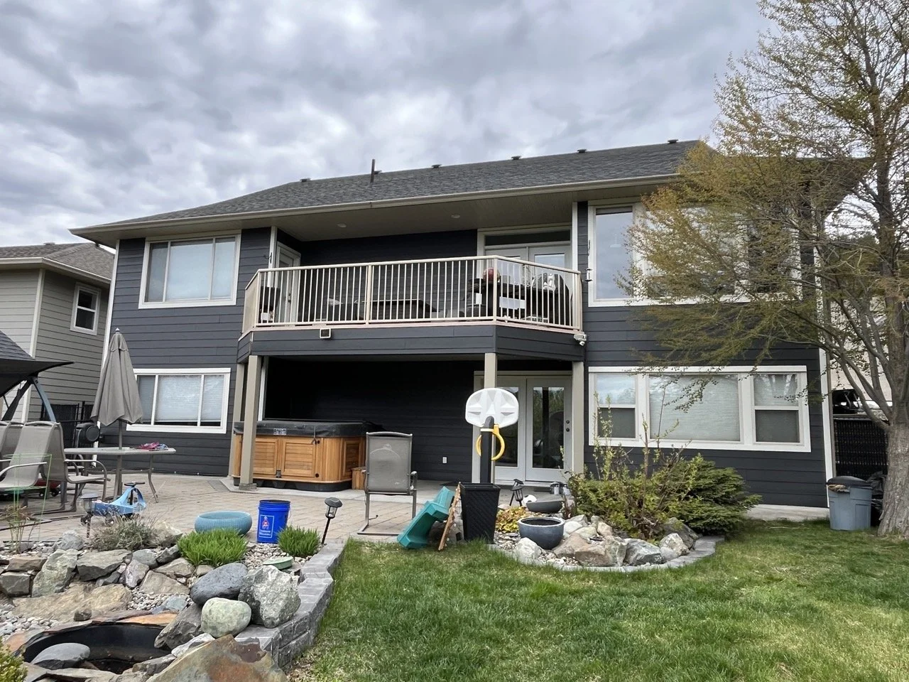 A two-story house with dark gray siding and white trim, featuring a balcony with a railing, patio furniture, a hot tub, and a well-maintained backyard with rocks, grass, and a tree.