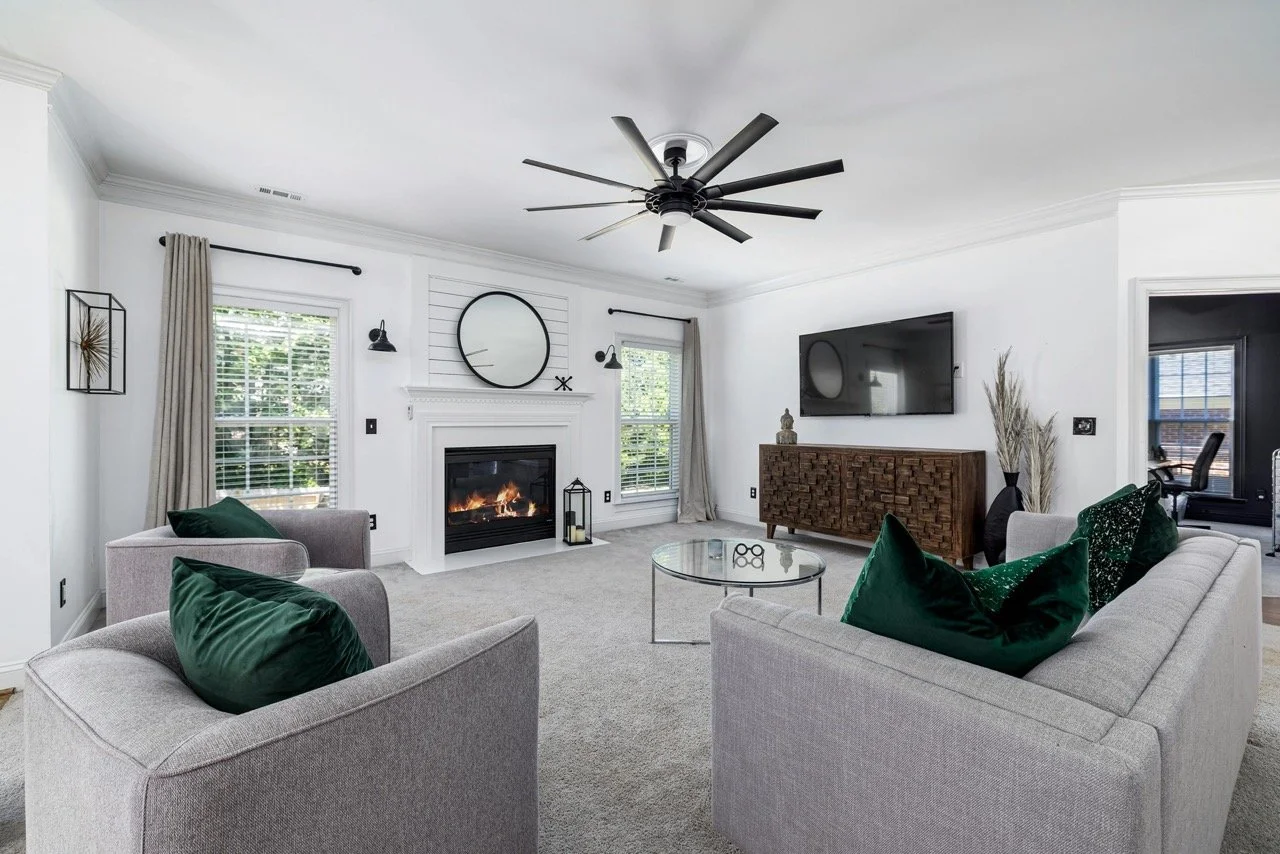Living room with gray sofas, green pillows, white walls, a fireplace, a wall-mounted TV, a wooden console, a round glass coffee table, black ceiling fan, and windows with beige curtains.
