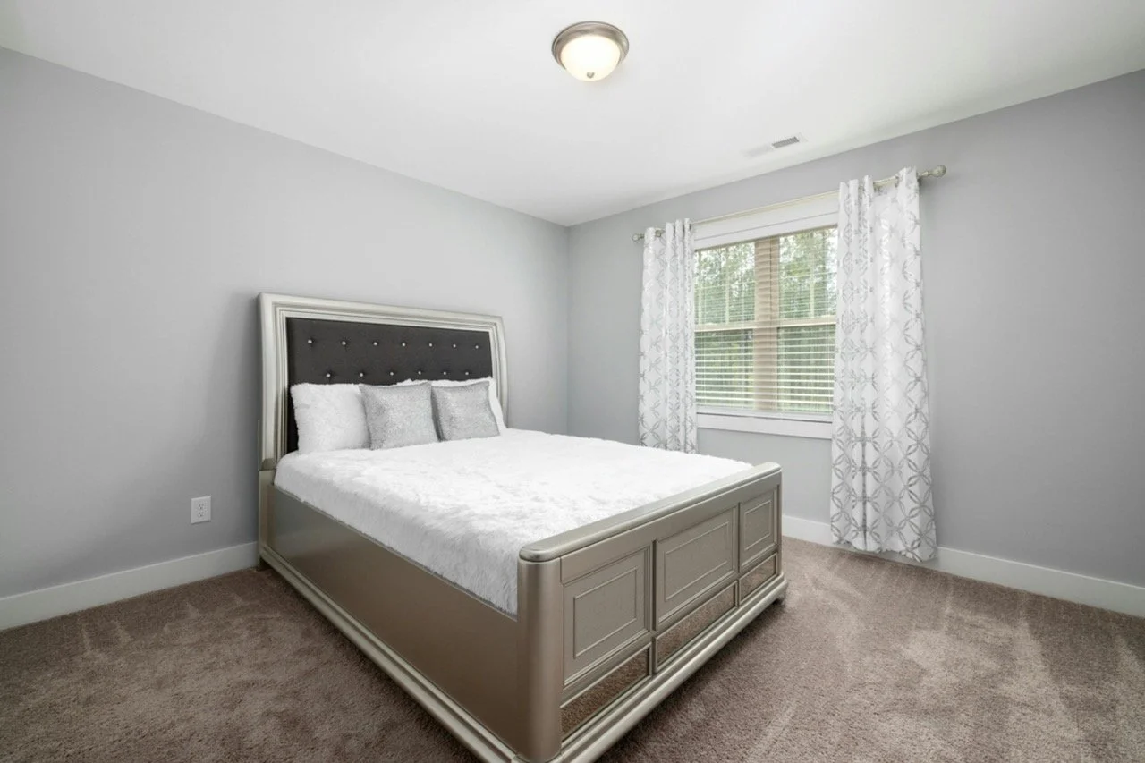 A bedroom with a bed, gray wall, window with white curtains, and beige carpet.