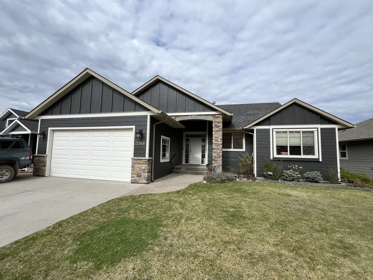 Photo of a modern single-story house with a black exterior, white garage door, and a front yard with grass and shrubs. The house features stone accents around the entryway and windows, with a cloudy sky overhead.