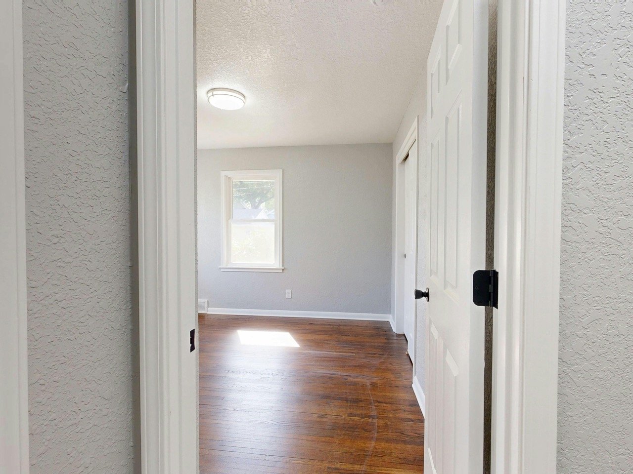 Empty room with white walls, hardwood flooring, a window, and a ceiling light, seen through a doorway.