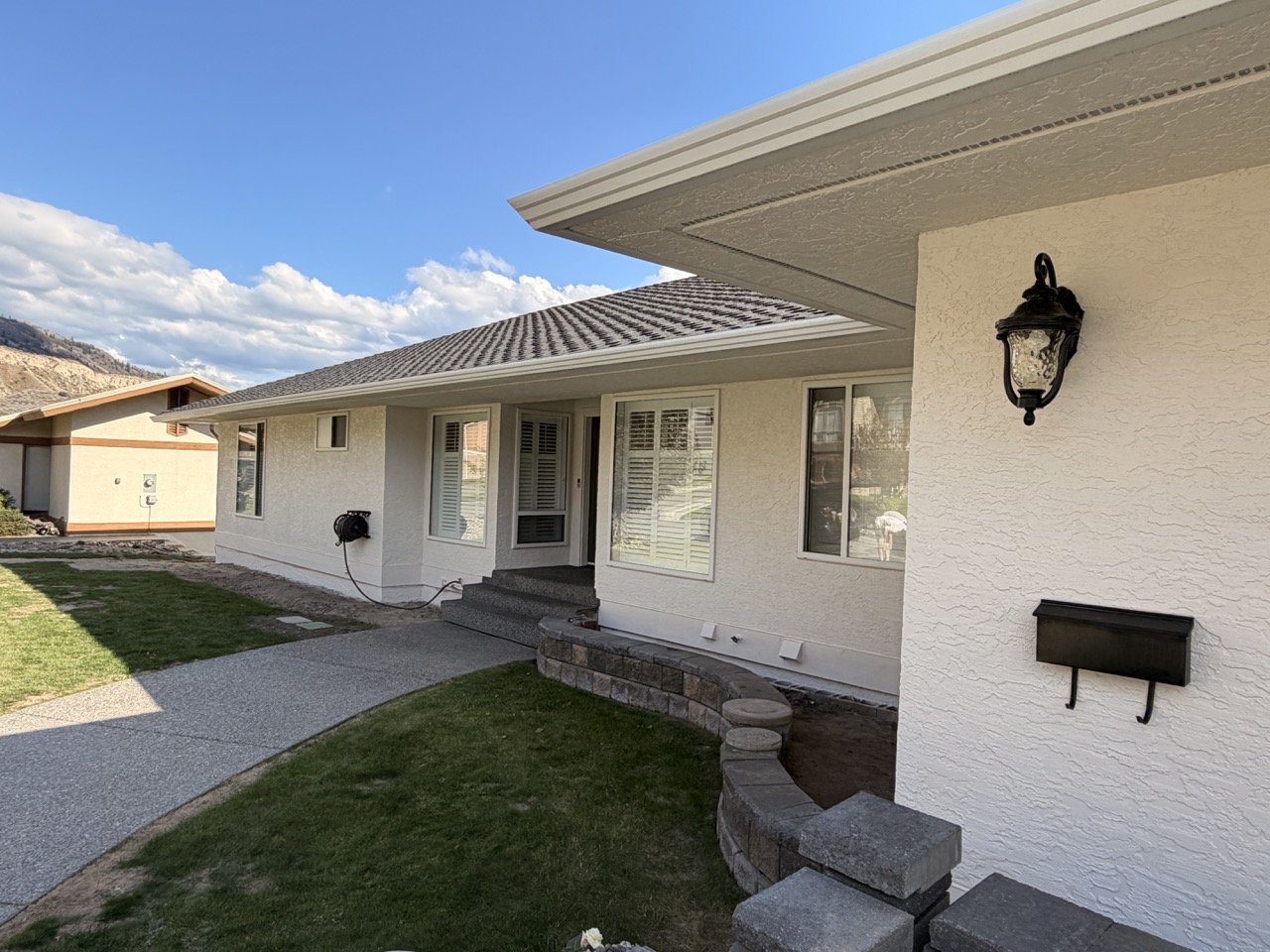 Photo of the front yard and house exterior, including a walkway, a mailbox, outdoor lighting, windows with shutters, and a garden with grass and concrete edging, under a partly cloudy sky.