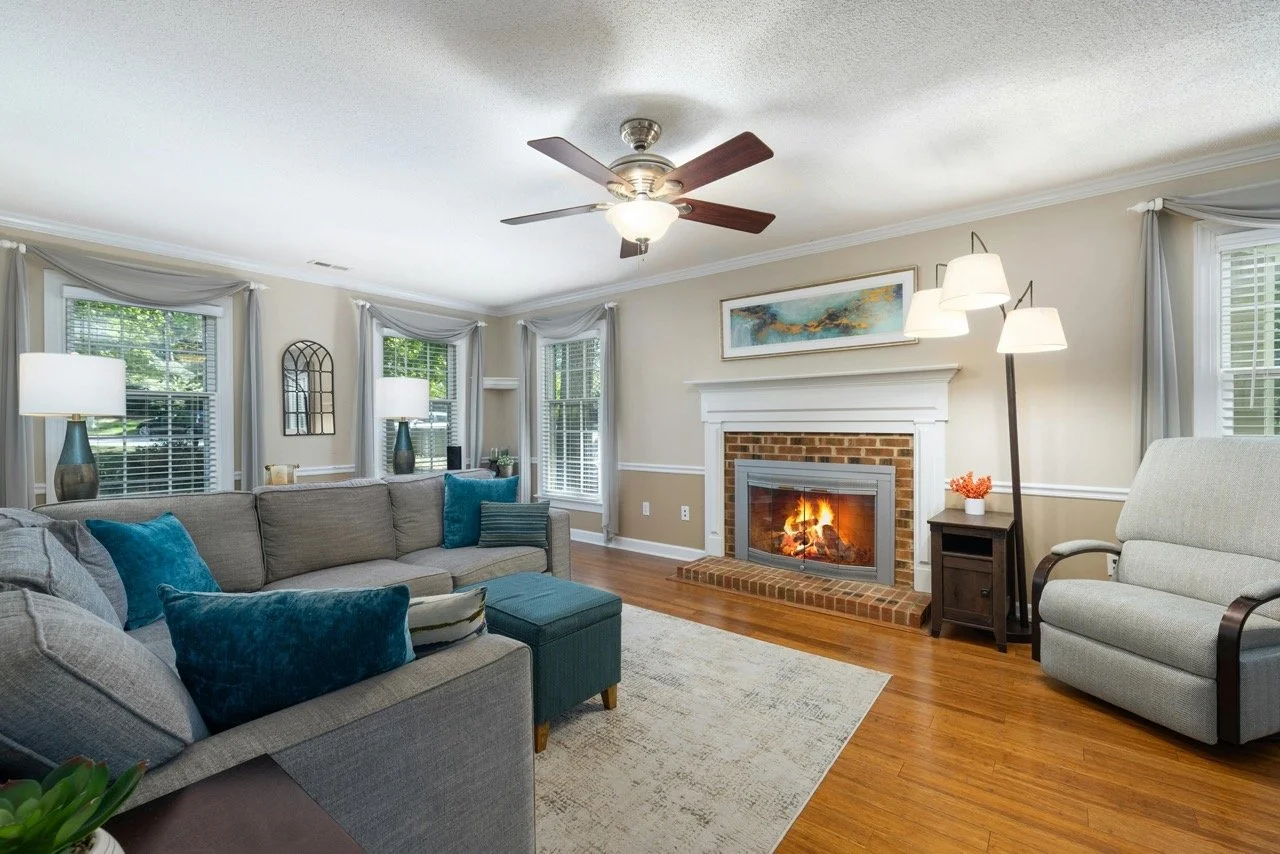 Living room with a fireplace, sofa with blue pillows, armchair, hardwood flooring, ceiling fan, and windows with curtains.
