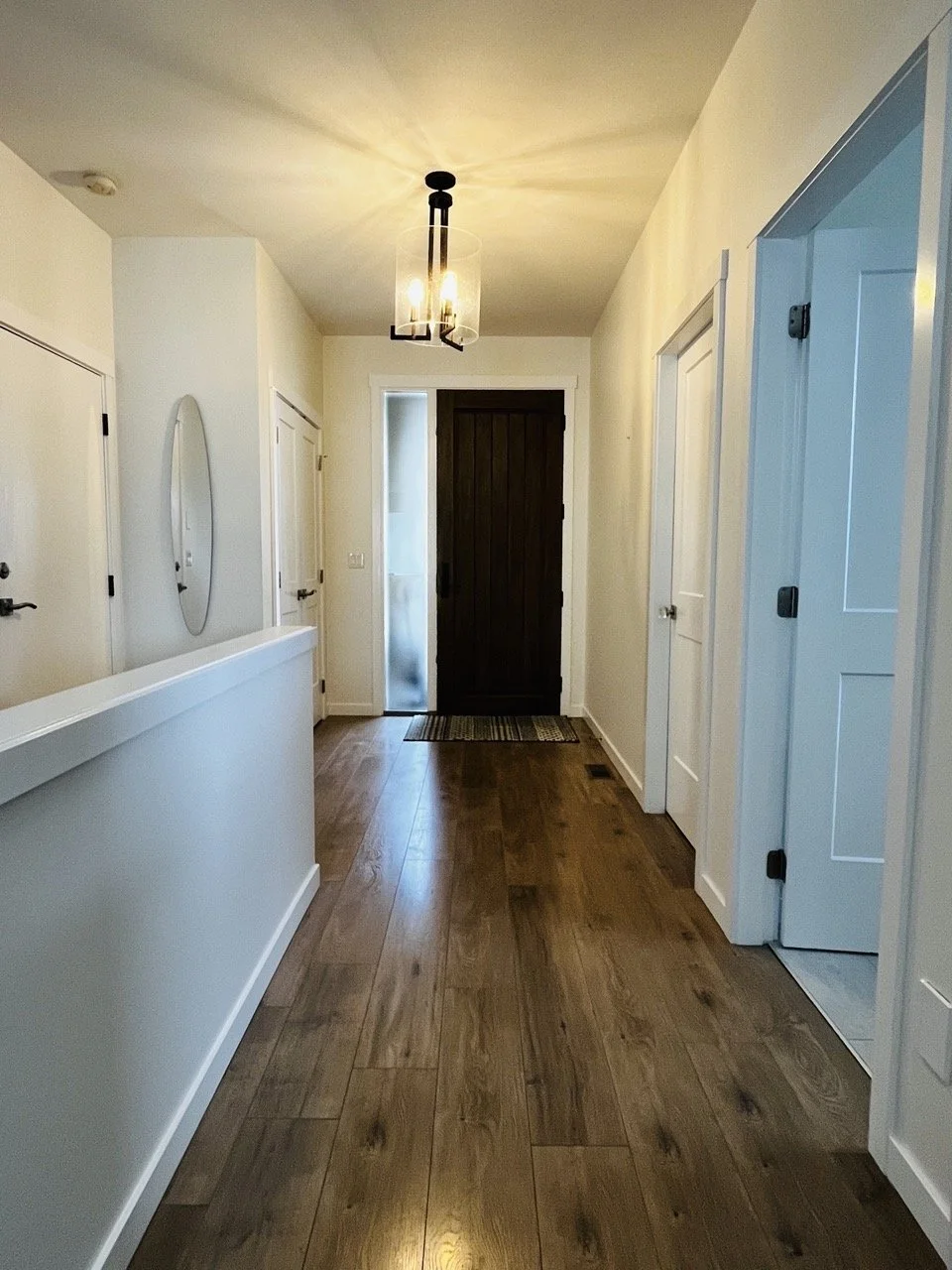 Interior hallway with hardwood floors, white walls, a dark front door with a frosted glass panel, and doorways on both sides. A modern ceiling light fixture is mounted on the ceiling.